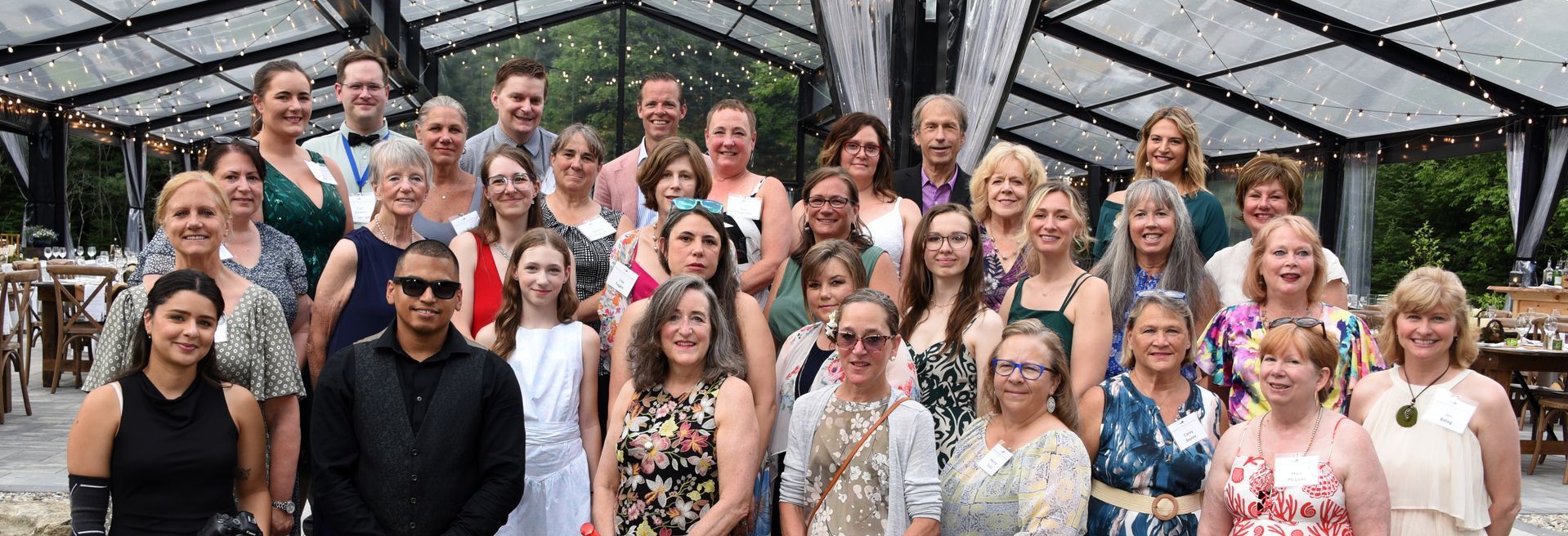 Group of people posing for a photo under a structure with a glass roof. Many are smiling, some wearing floral dresses.