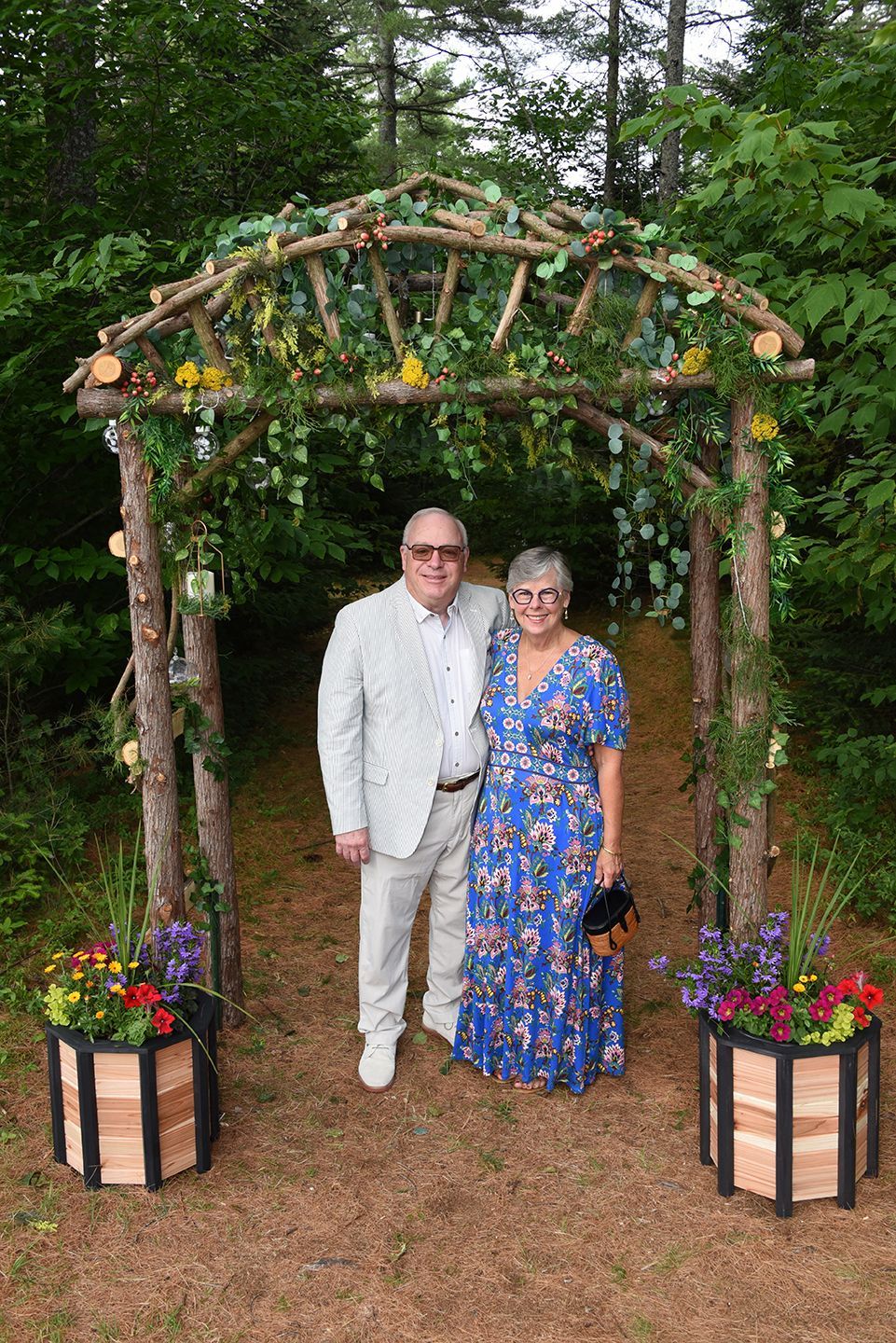 Couple smiling, posing under a wooden archway decorated with vines and flowers in a natural setting.