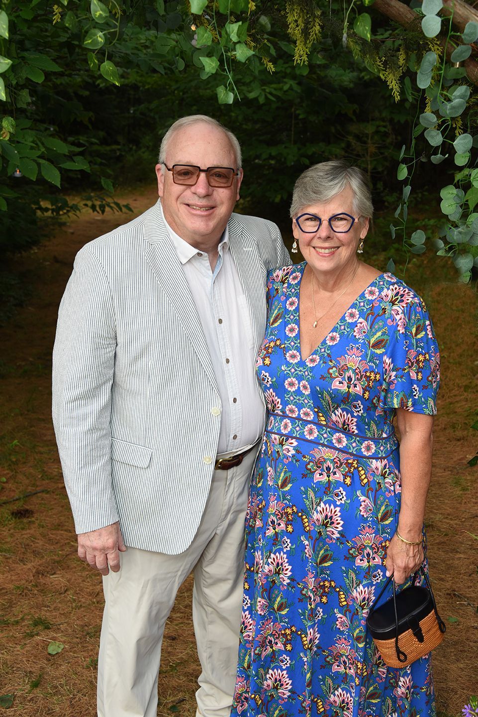 Older couple posing outdoors. Man in seersucker jacket, woman in blue floral dress, both smiling.