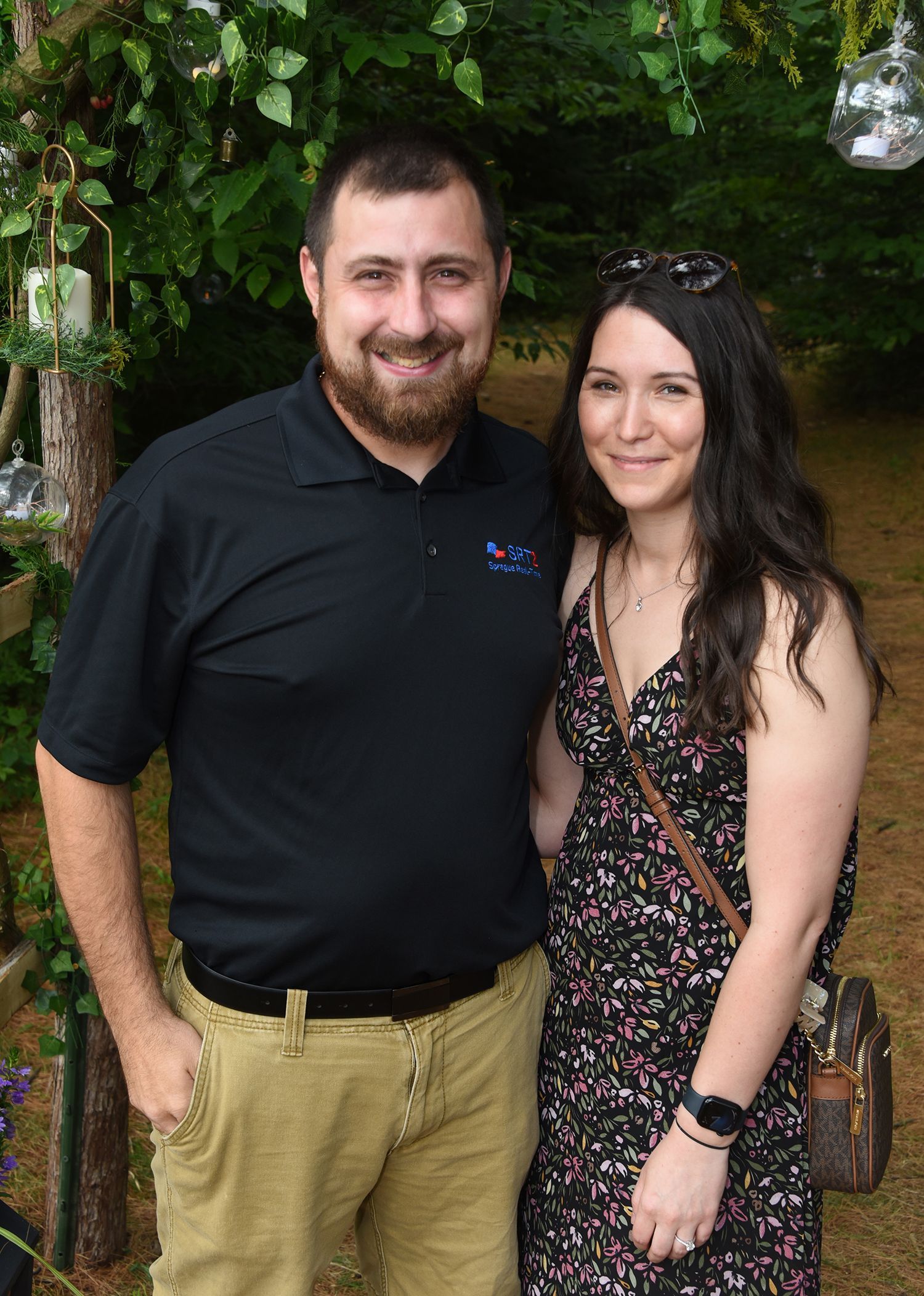 A man with a beard and a woman in a floral dress pose together outdoors. They are both smiling.