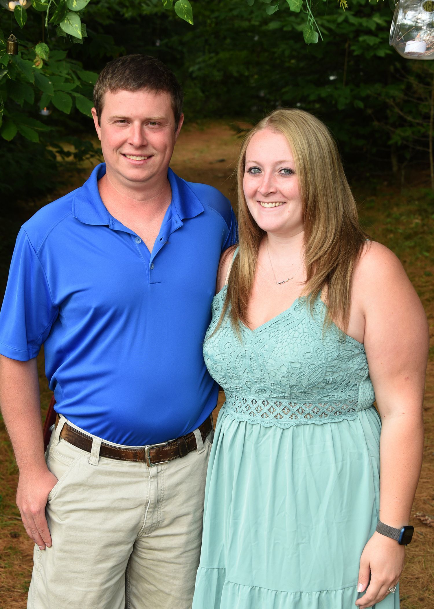 A man and woman stand close together outdoors. The man wears a blue shirt and khakis. The woman wears a teal dress.