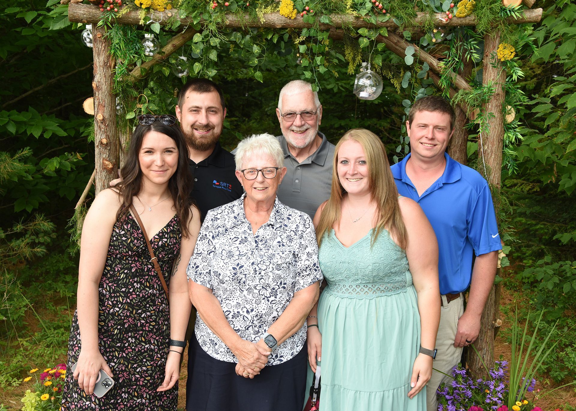 Family posing under a floral archway. Six people smile, with the older woman in the front. Lush greenery in the background.