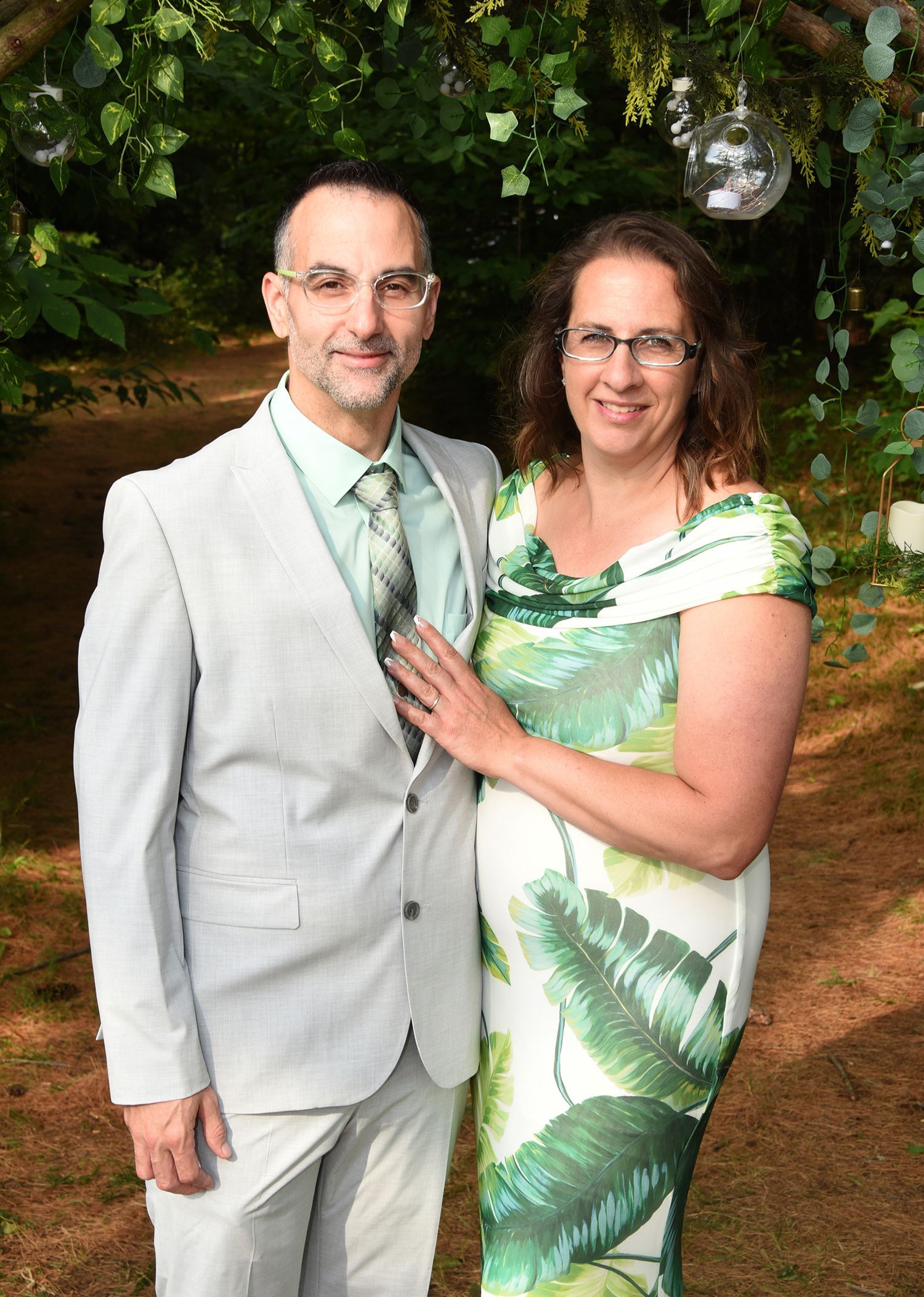 Couple posing outdoors. The man wears a light suit, the woman a green tropical print dress. They are smiling.