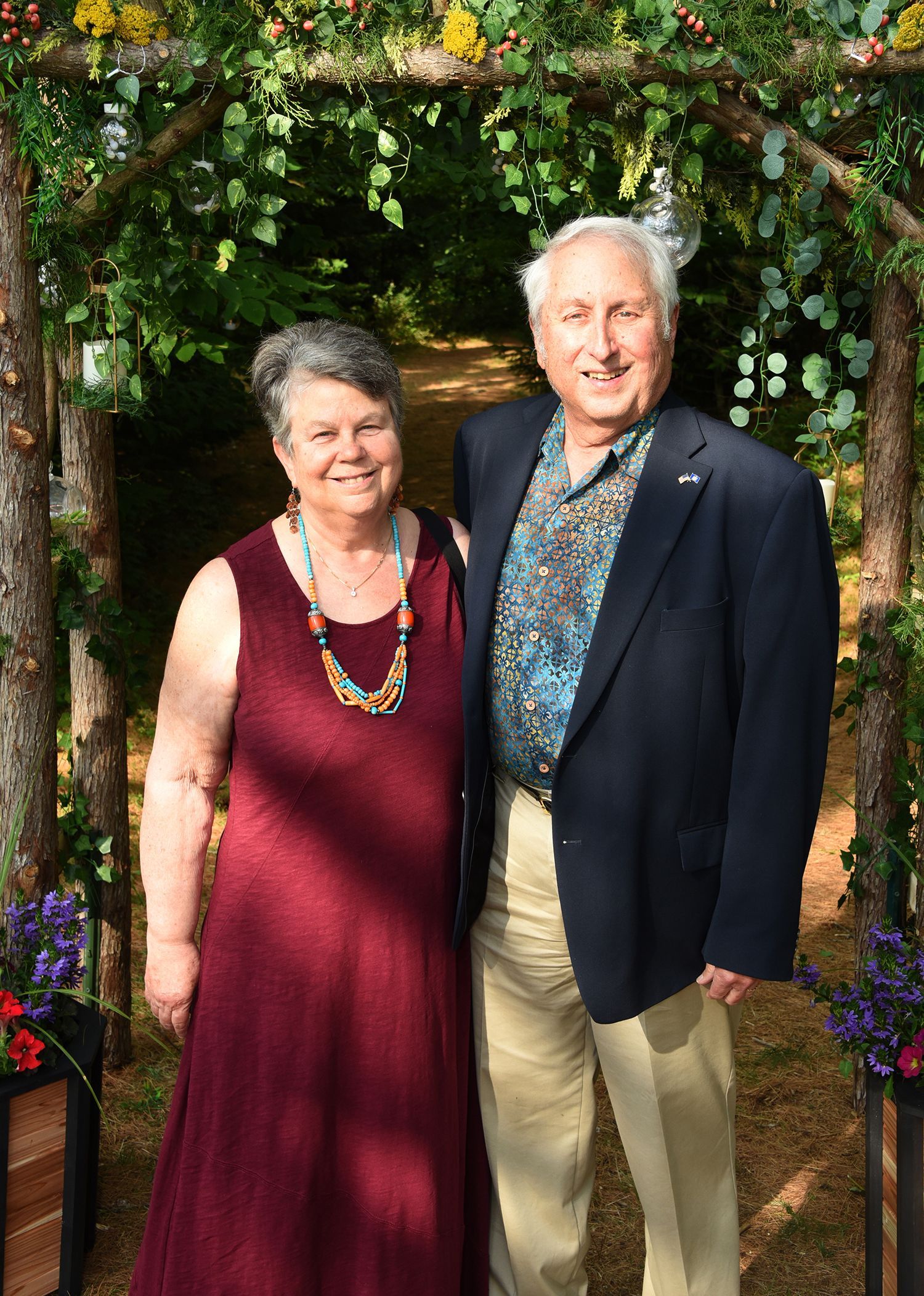 An older couple stands together under a flower-adorned archway. 