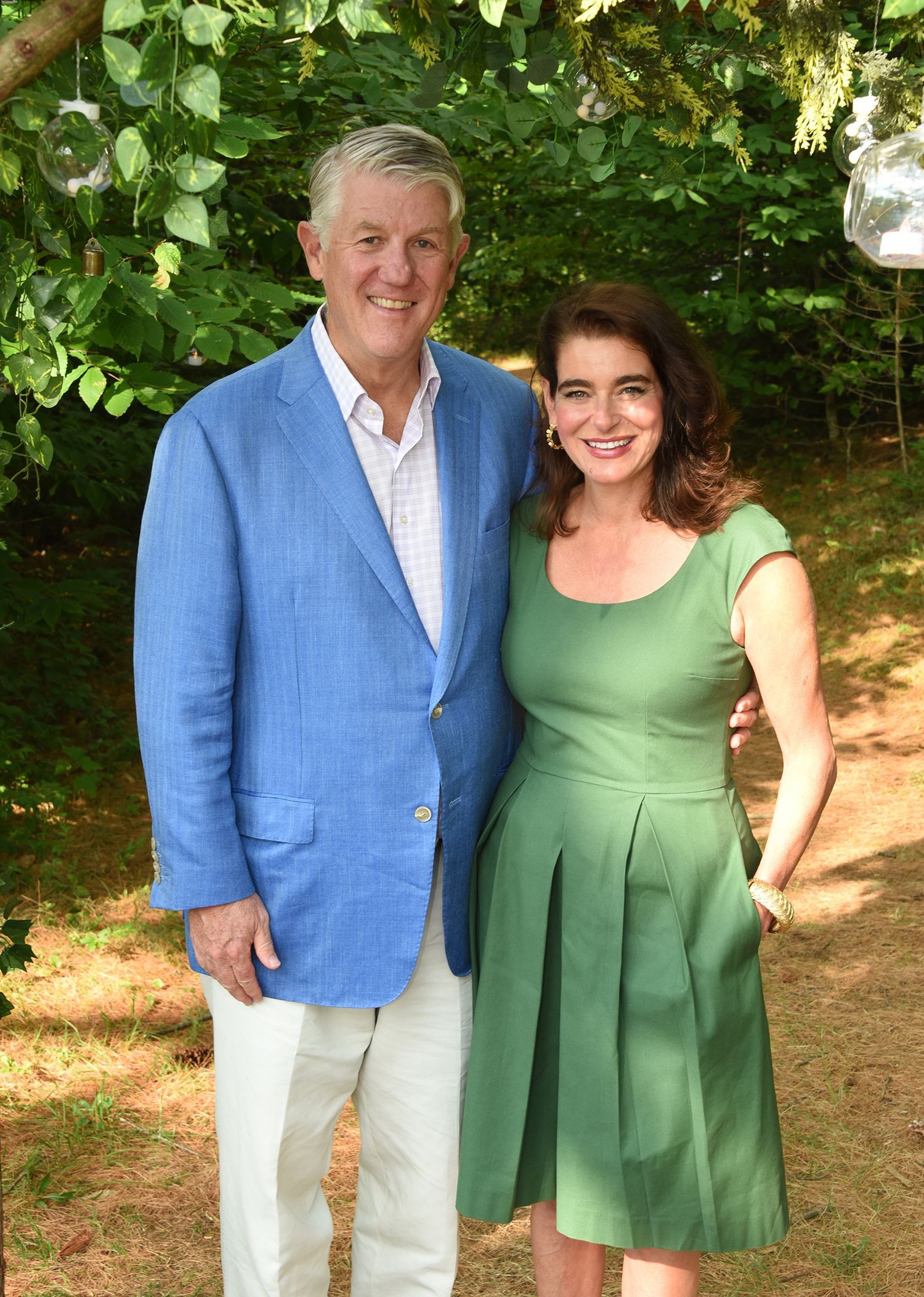 Man in blue blazer and woman in green dress smiling together outdoors. Lush green foliage in the background.