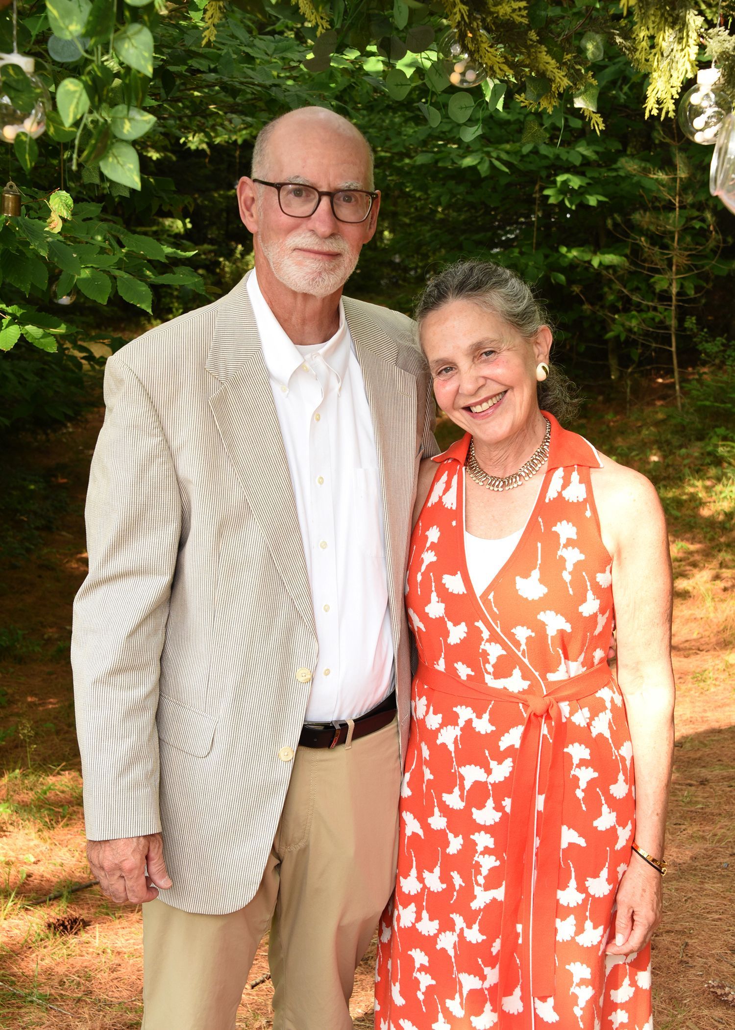 A smiling older couple poses outdoors. The man wears a striped blazer, the woman a red floral dress.