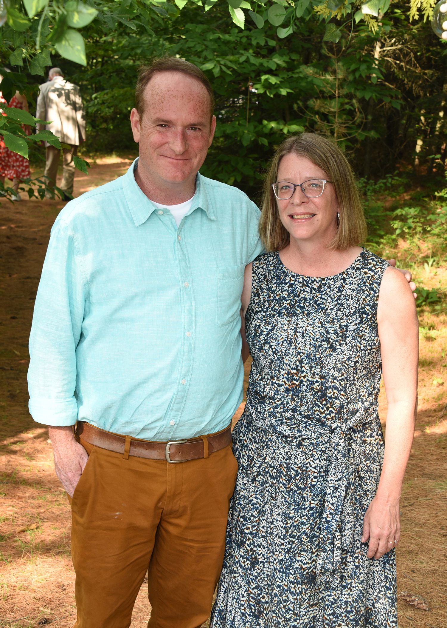 A man and woman stand outdoors. The man wears a blue shirt and brown pants. The woman wears a patterned dress and glasses.
