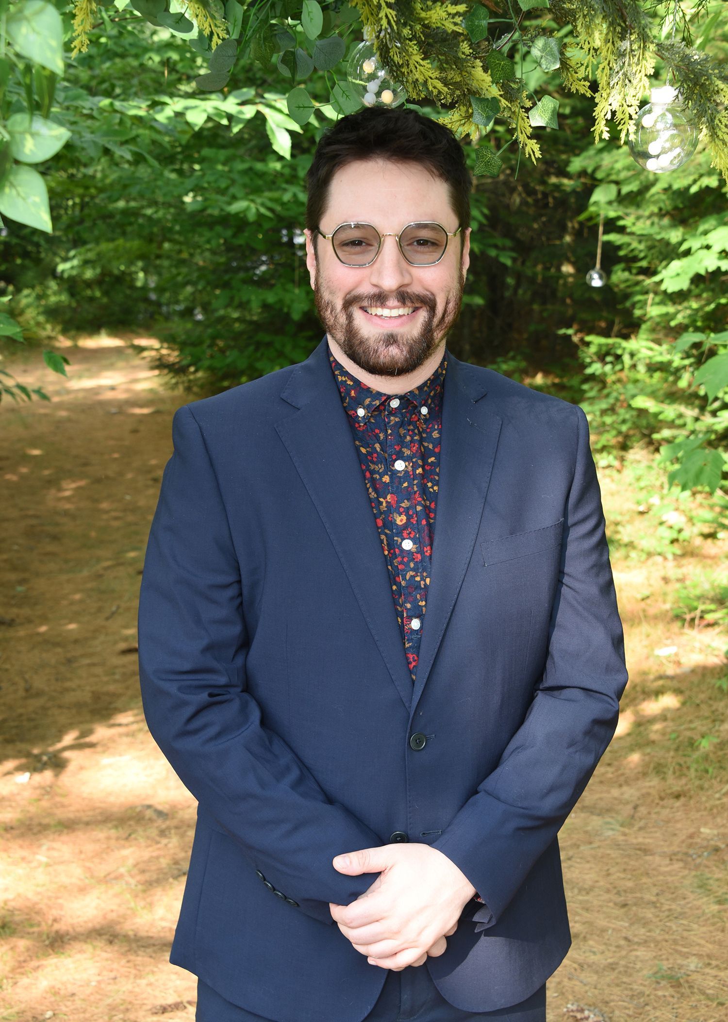 A man with glasses and a beard wearing a blue suit over a patterned shirt, standing outdoors in a wooded area.