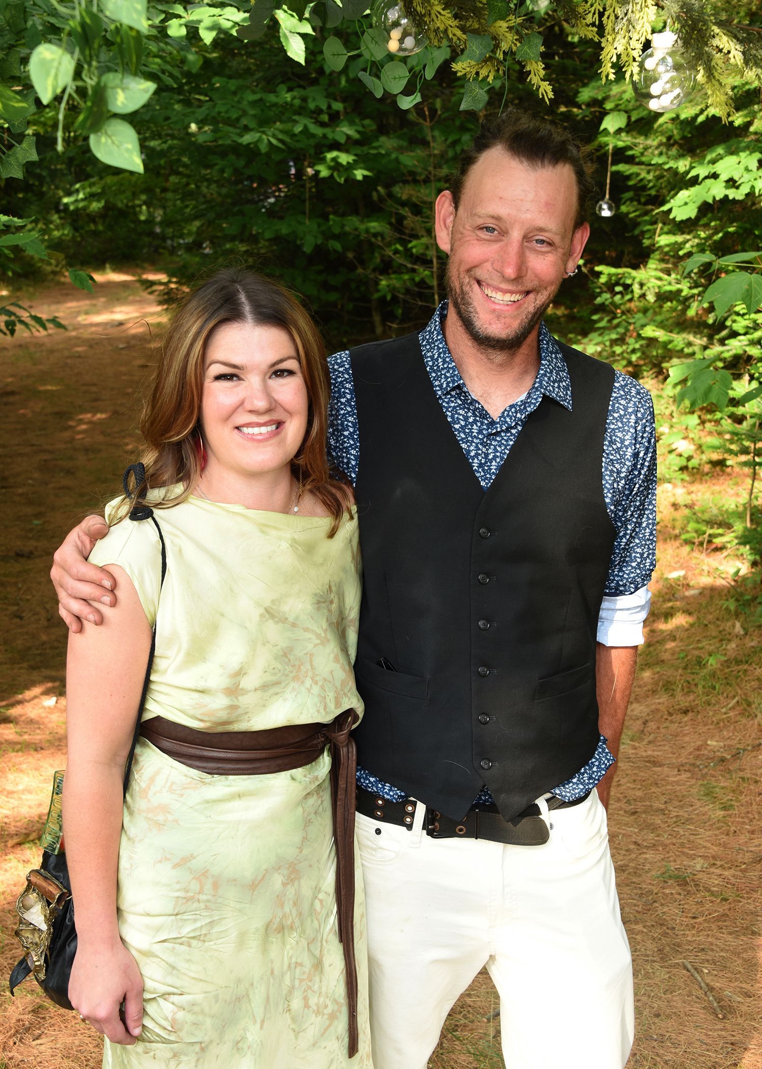 Woman in light green dress and man in vest smiling, standing outdoors with trees in the background.