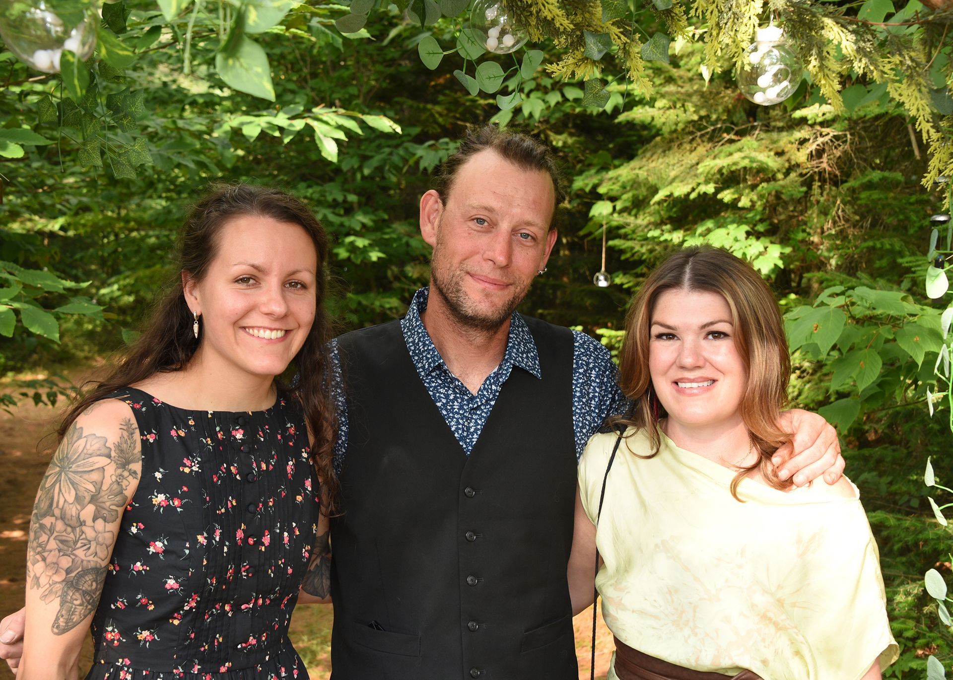 Three people posing outdoors. A man in a vest smiles between two women; trees and greenery are in the background.