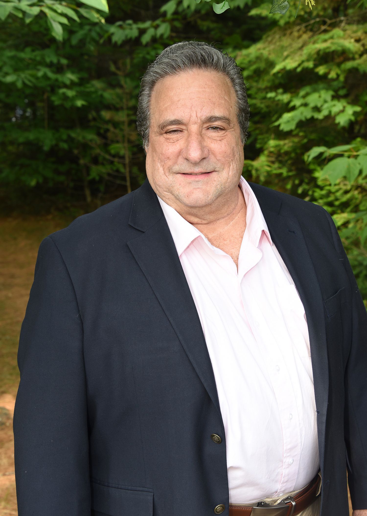 Man in a navy blazer and pink shirt smiles outdoors against a green backdrop.