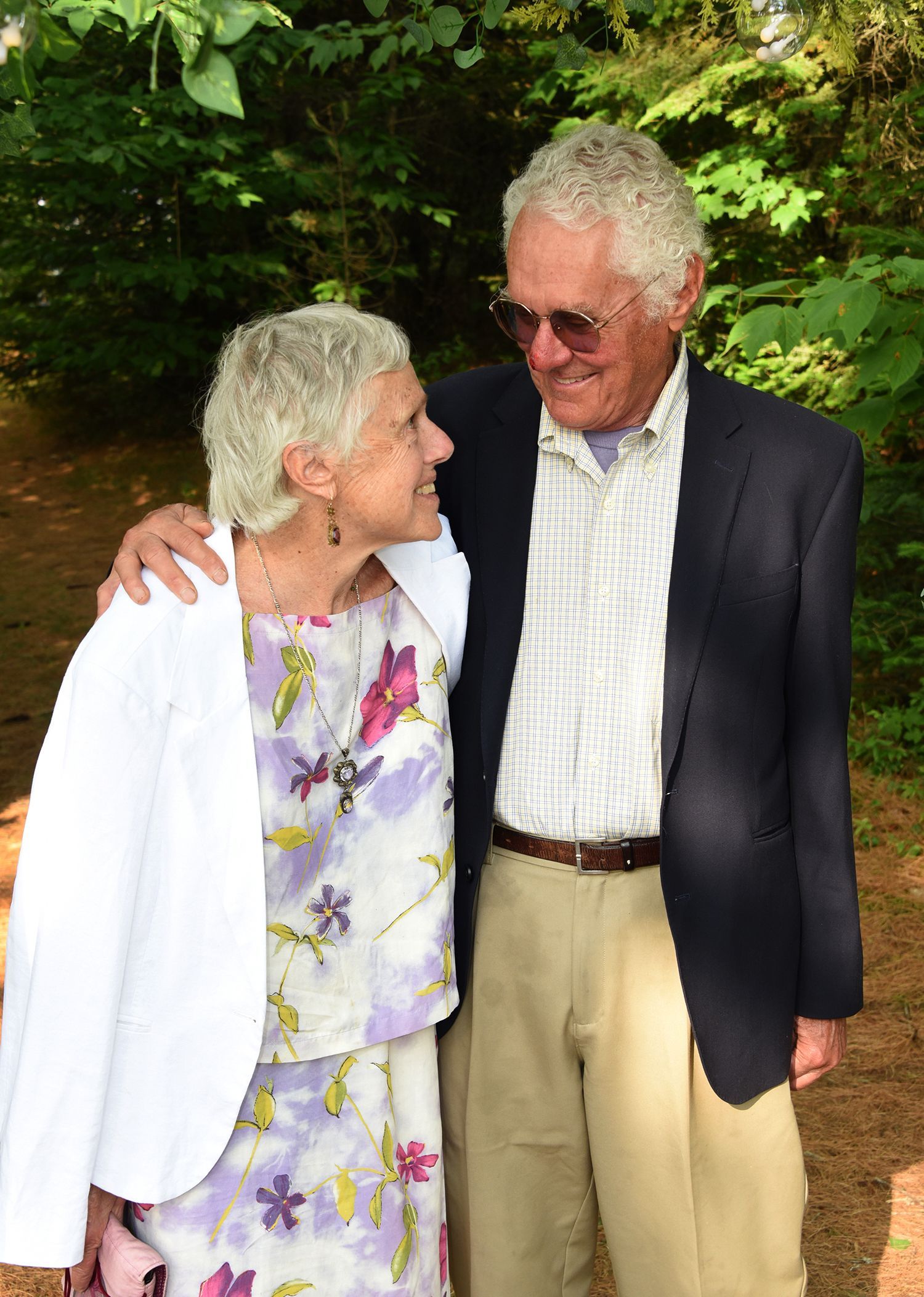 An elderly couple smiles at each other, standing outside. The man has his arm around the woman.