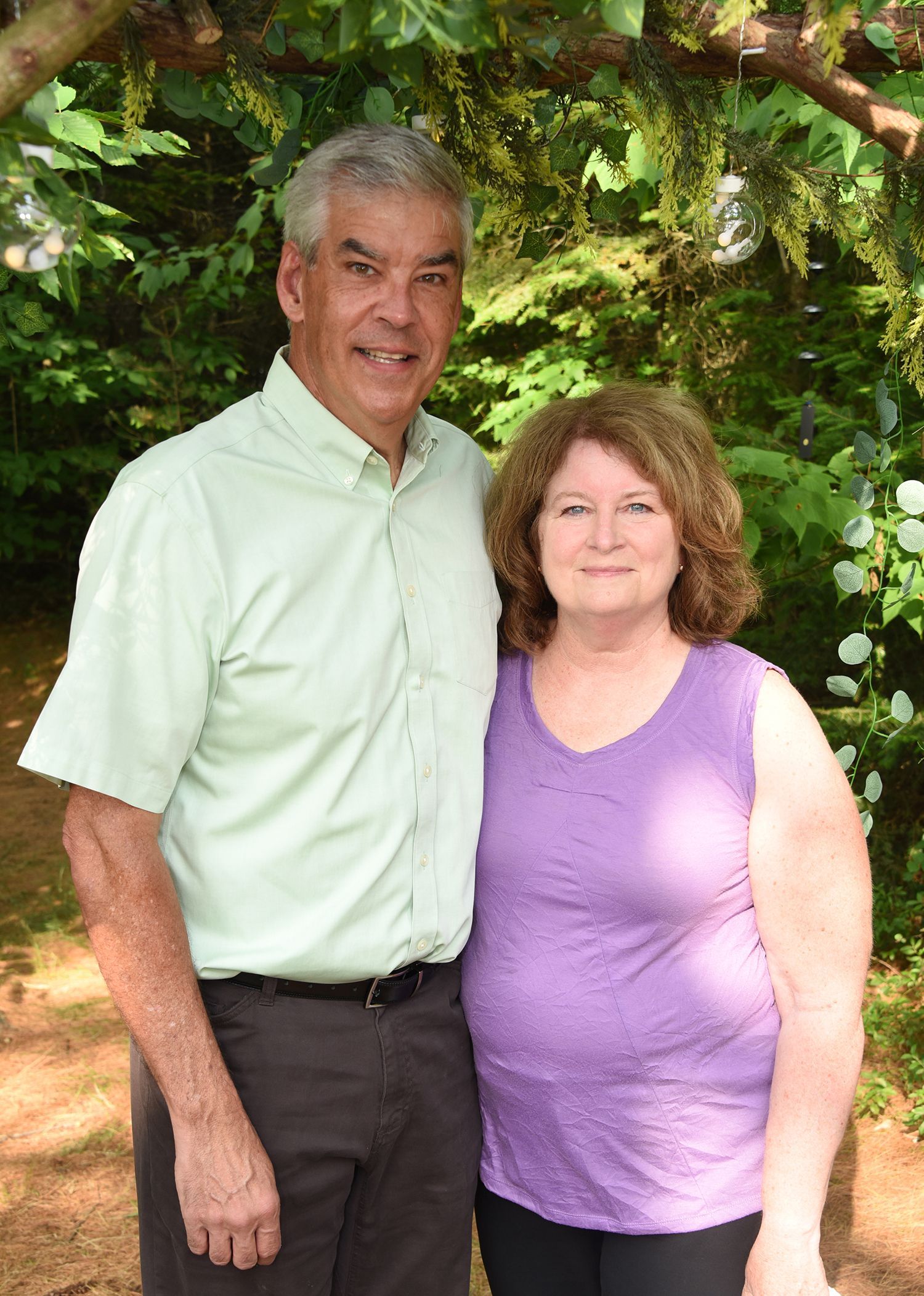 A smiling older man and woman stand closely together outdoors, with lush greenery in the background.
