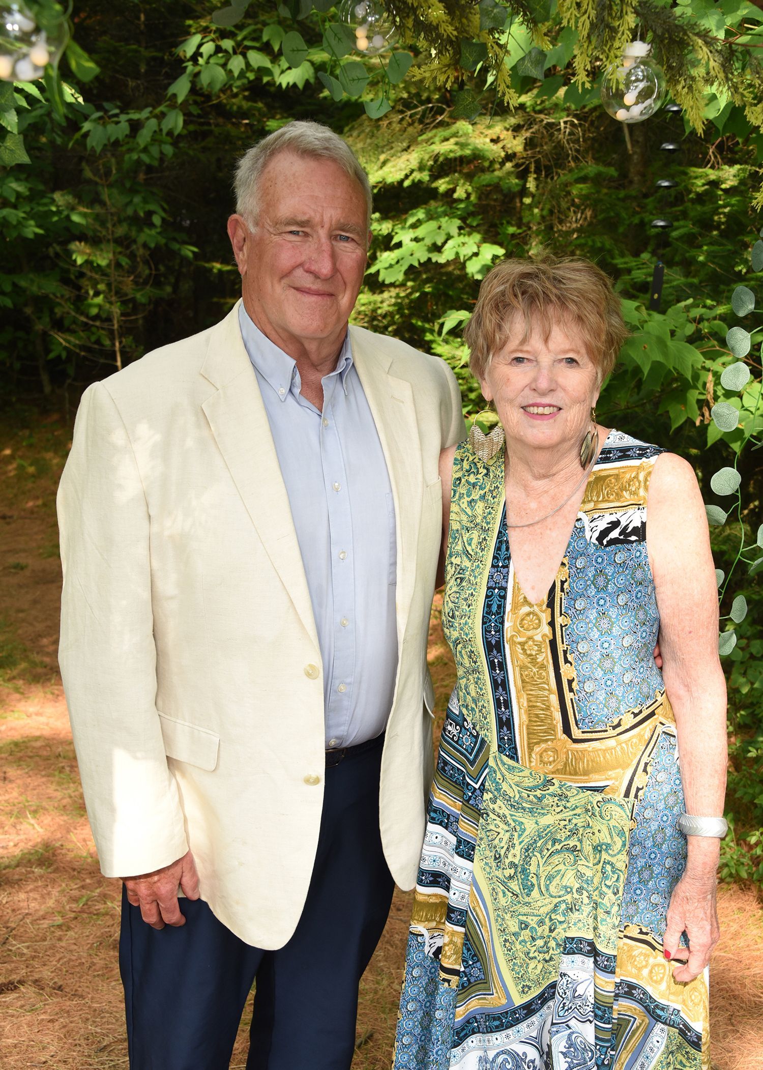 An older couple smiling and posing outdoors. The man wears a cream blazer, the woman a patterned dress.