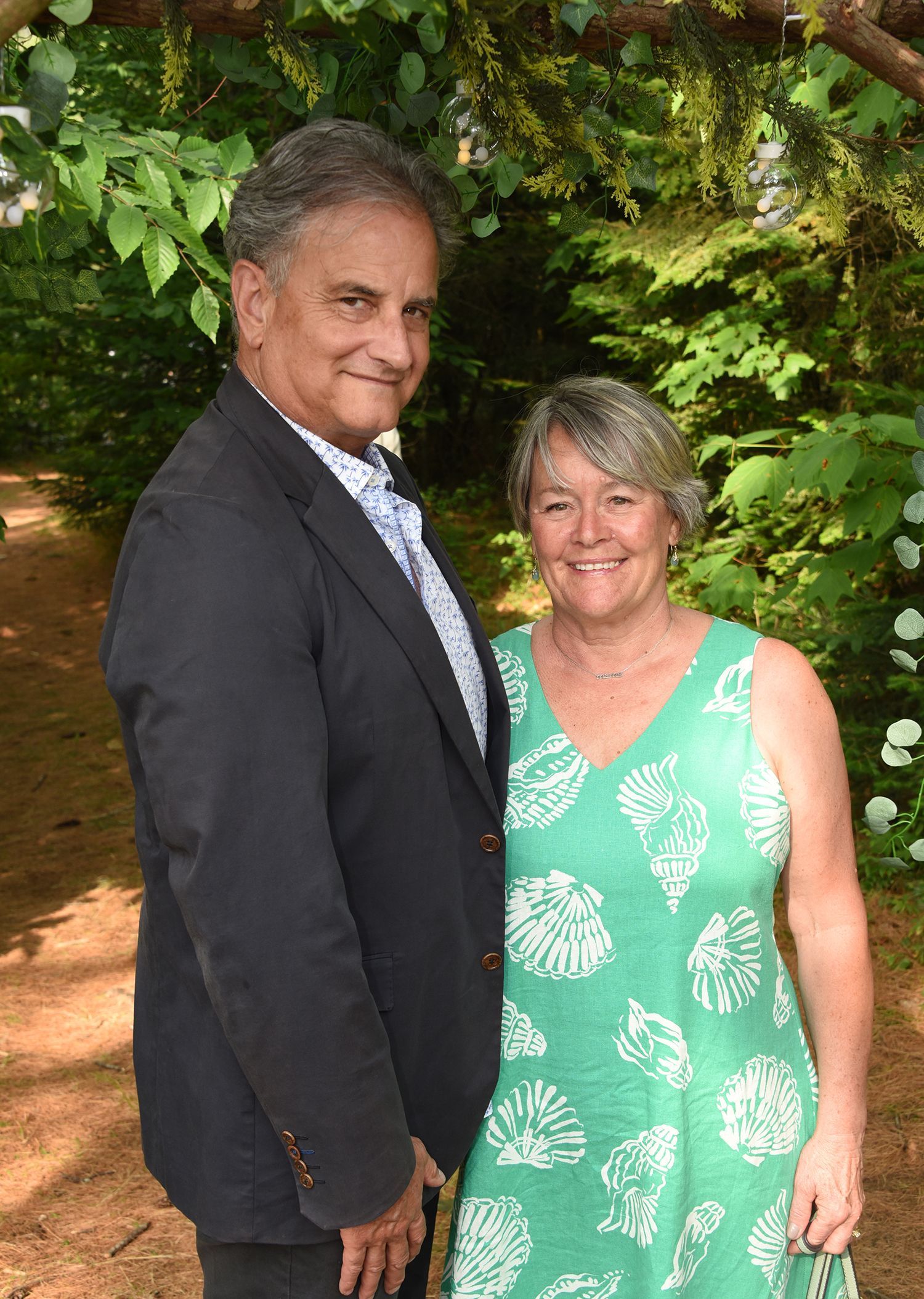 A smiling couple poses together outdoors in front of greenery; man in a blazer, woman in a green dress.