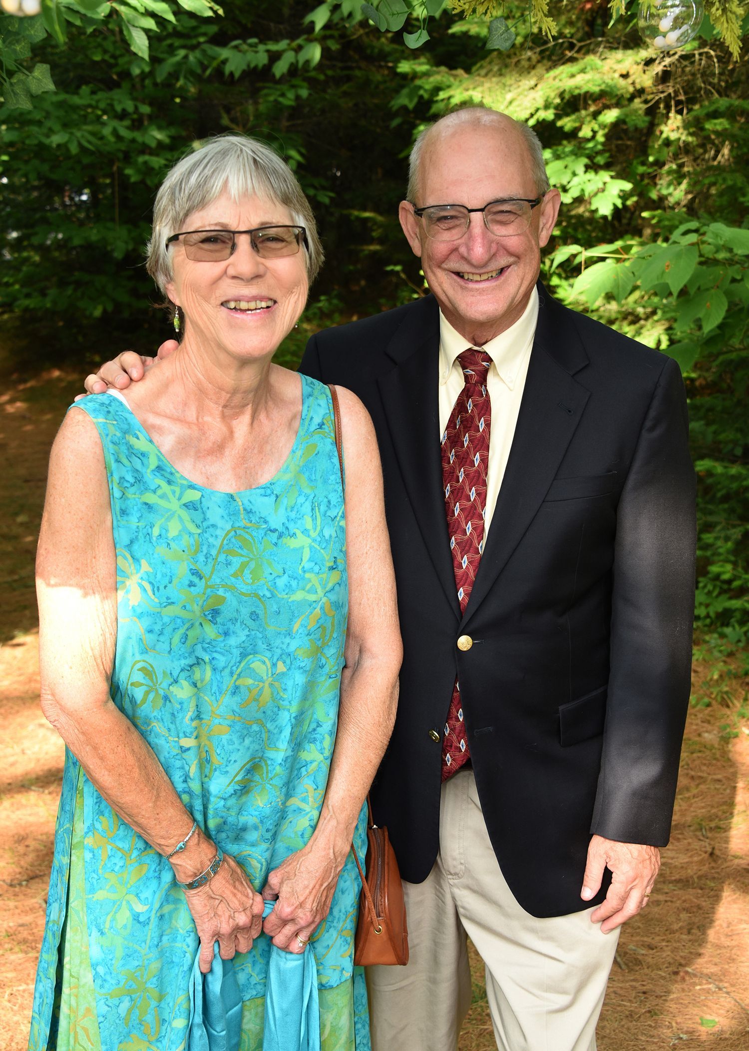 Smiling older couple posing outdoors. Woman wears a turquoise dress; man in blazer, tie.