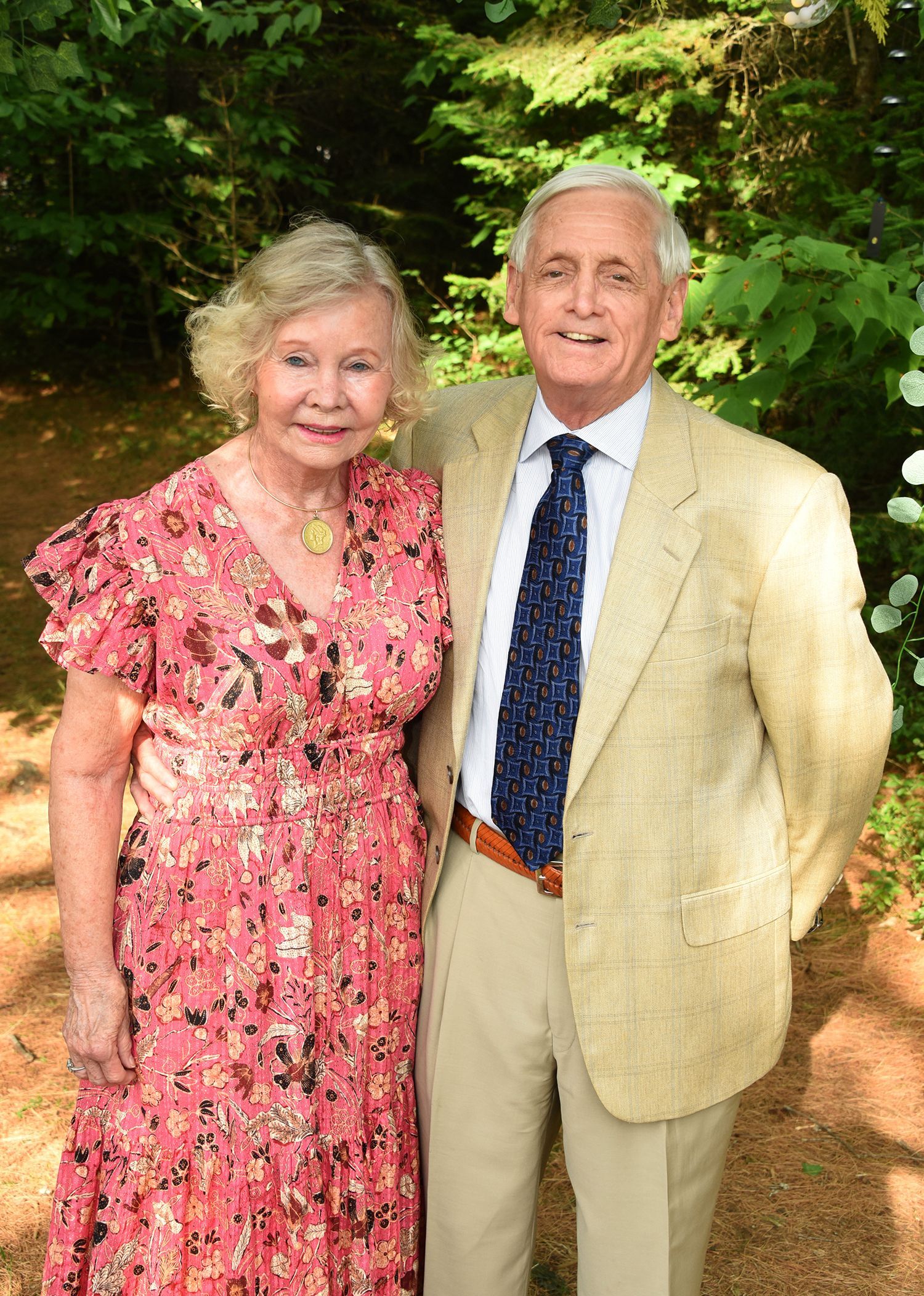 An elderly couple stands close, posing outdoors. The woman wears a pink floral dress, the man a tan blazer and patterned tie.