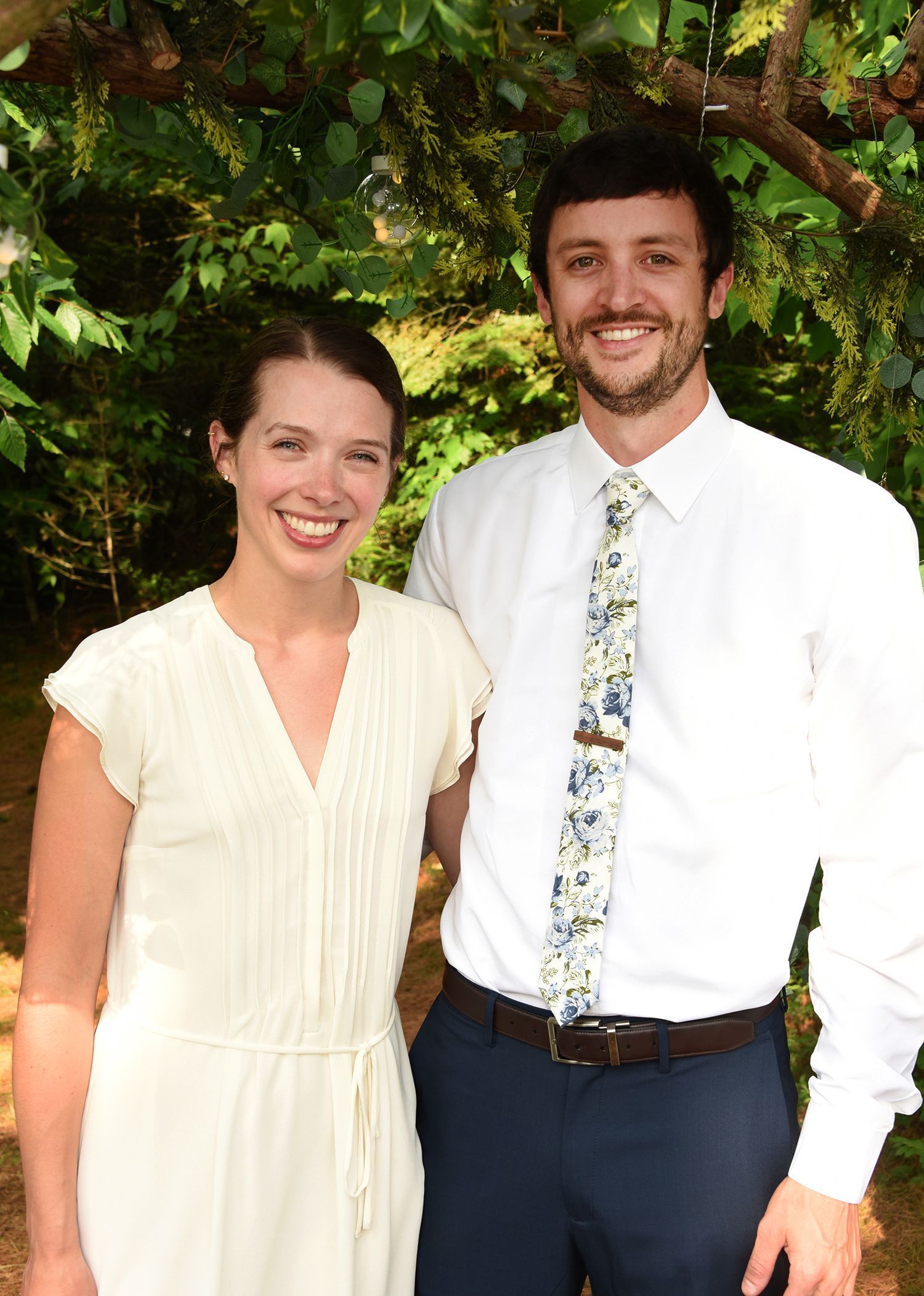 A smiling couple poses outdoors under a tree. The woman wears a cream dress; the man wears a white shirt and patterned tie.