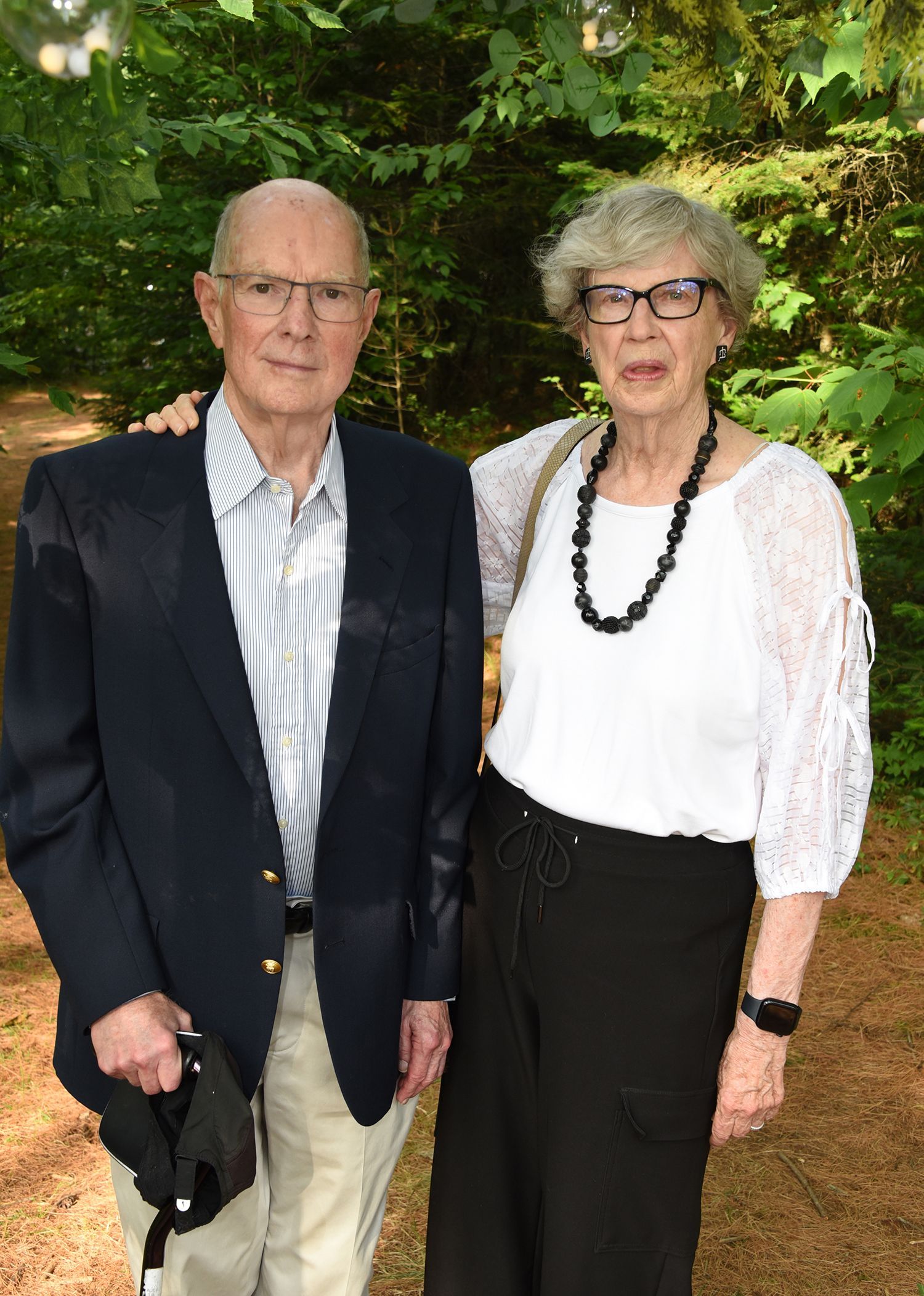 Elderly couple standing outdoors; the man in a blazer and the woman with a white top and black pants. 
