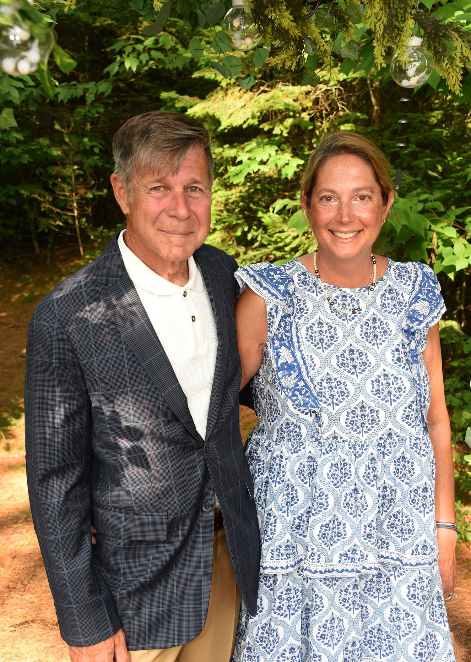 An older man with a blue patterned blazer and a woman in a blue and white dress pose together in front of greenery.