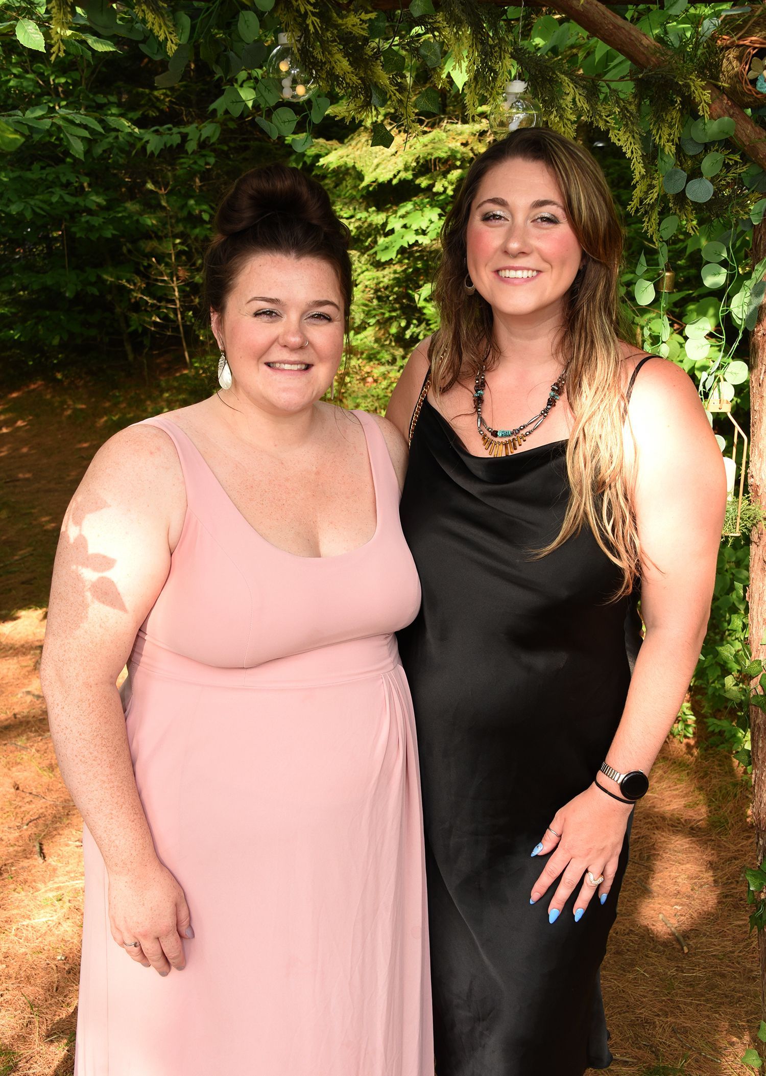 Two women pose outdoors. One wears a pink dress, the other a black slip dress with a necklace. 