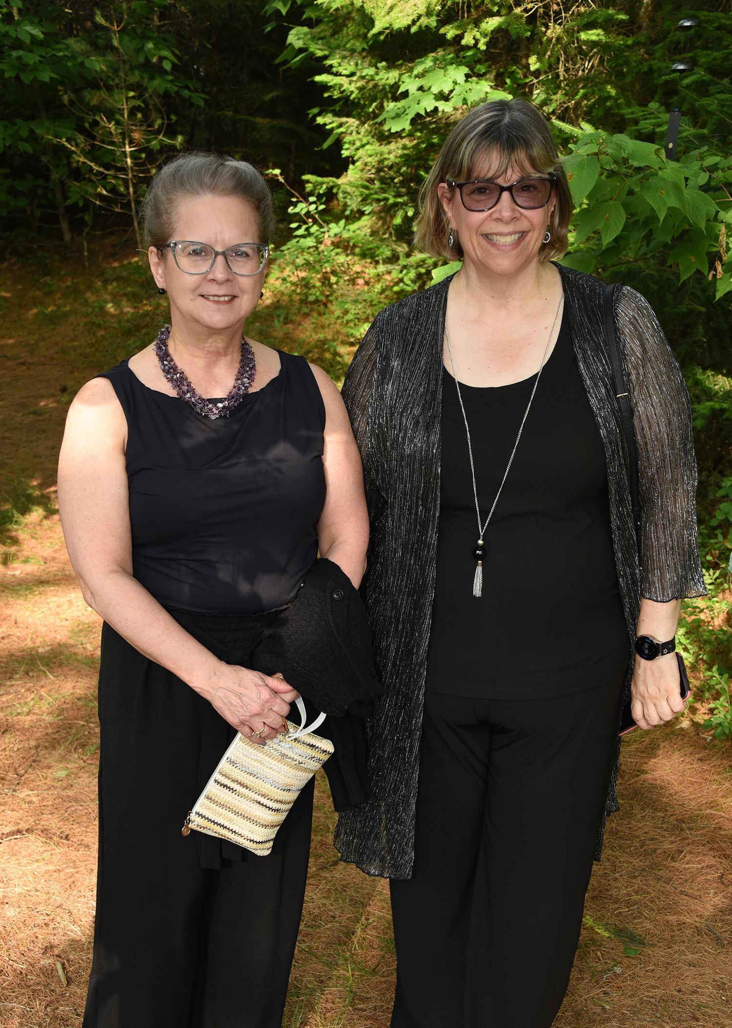 Two women in black outfits stand outdoors by trees. One holds a clutch purse. Both wear glasses.