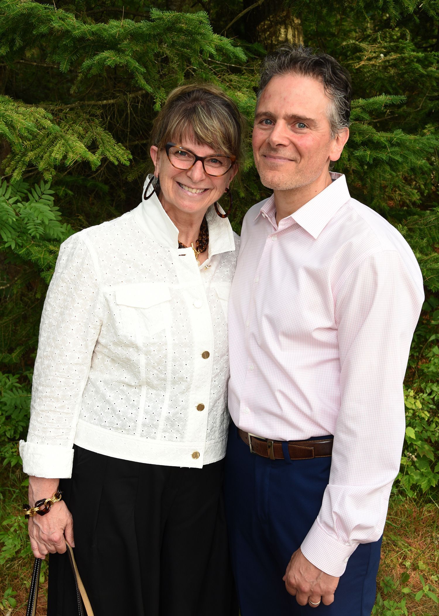 A smiling man and woman pose together outdoors. The woman wears a white jacket and glasses; the man wears a pink shirt.