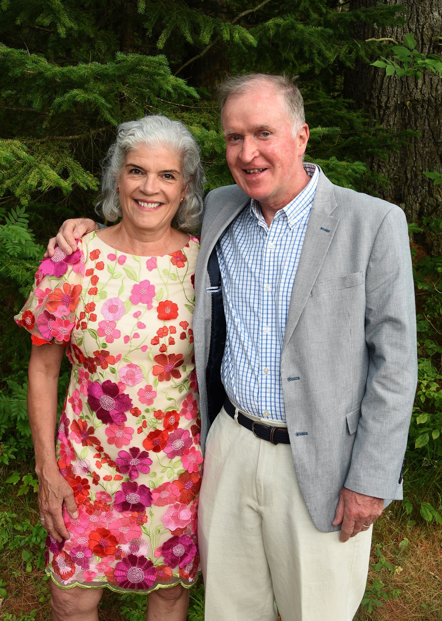 A smiling older couple poses outdoors. The woman wears a floral dress, and the man wears a blazer and light pants.