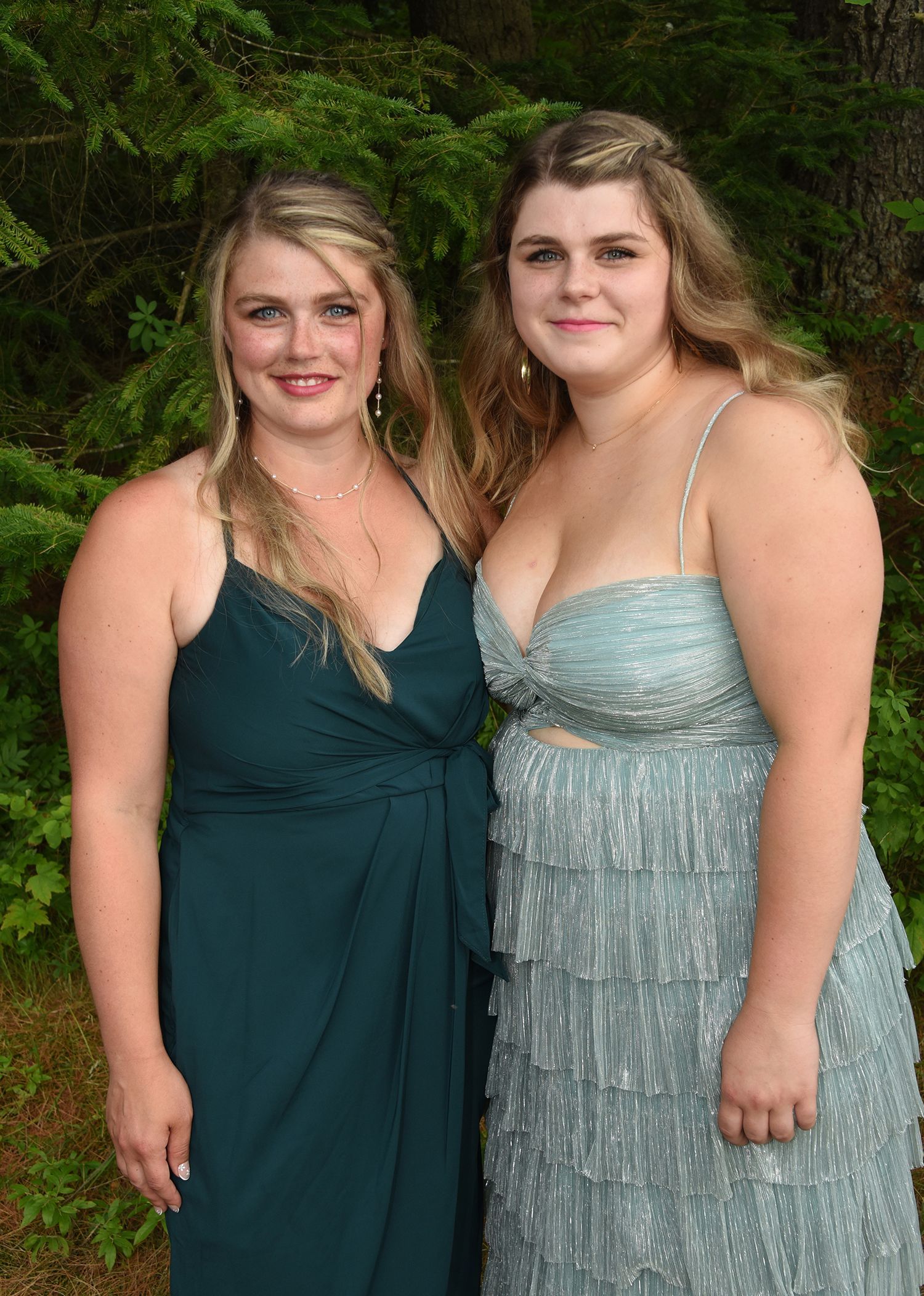 Two young women in formal dresses pose outdoors. One wears teal, the other light blue with layered fringe.