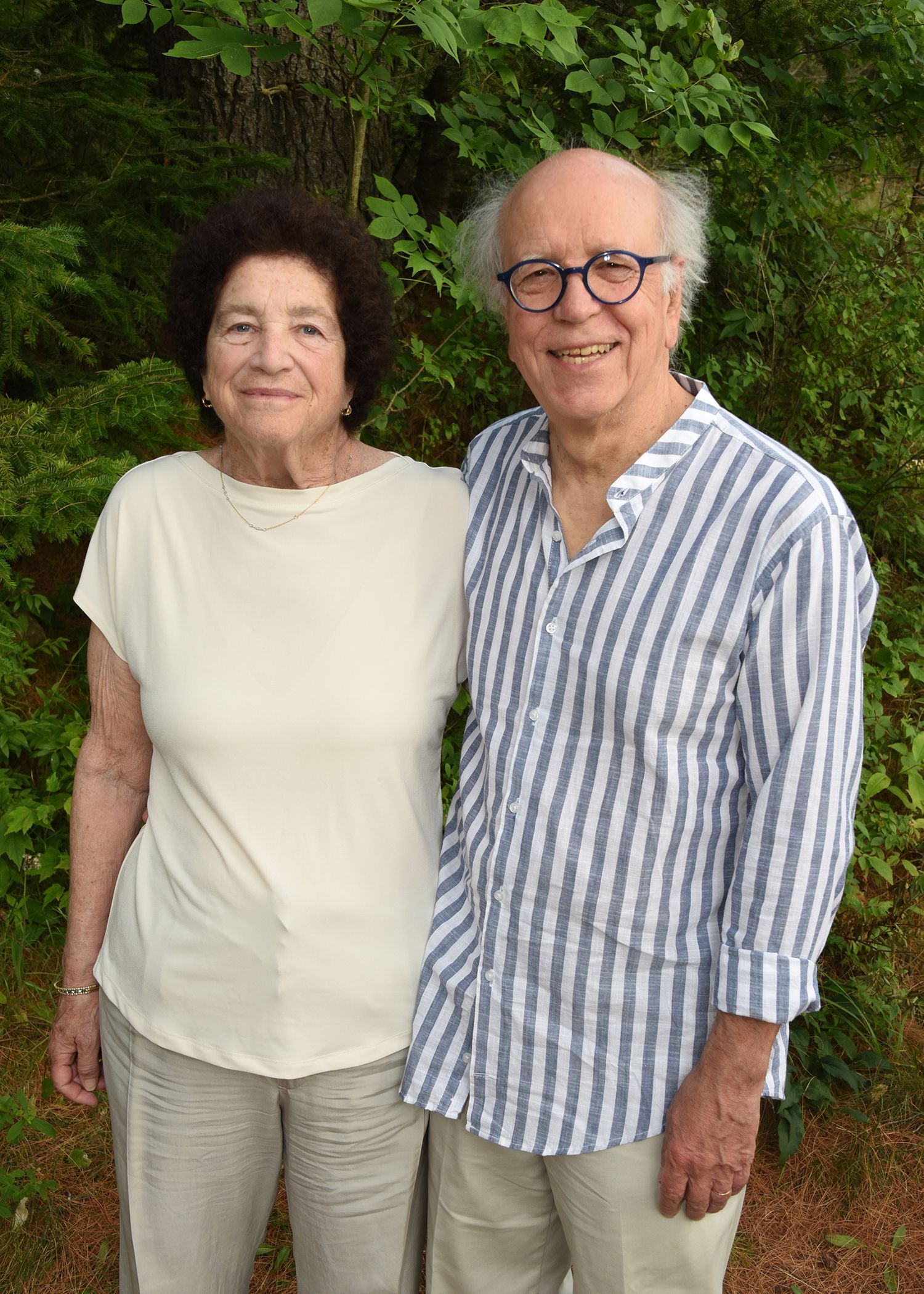 An elderly couple, a woman and a man, stand outdoors smiling. The woman wears a cream shirt and pants.