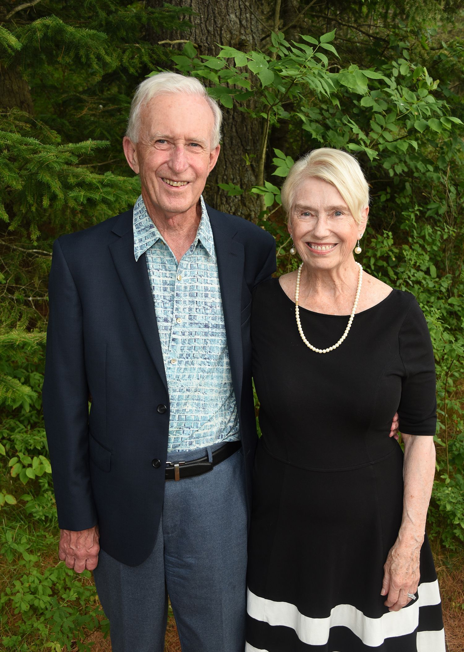 An elderly couple smiling at the camera. The man wears a navy blazer, blue shirt, and jeans. 
