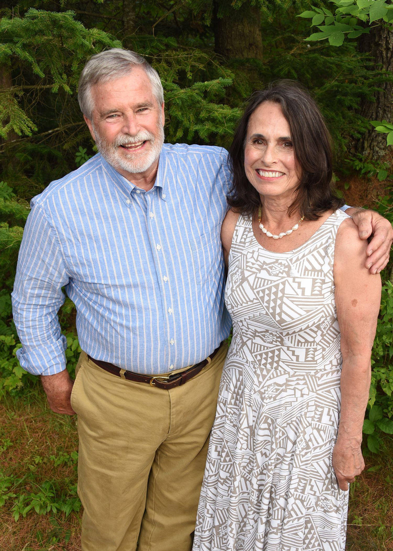 An older couple smiles and poses outdoors. The man has a beard, wearing a blue shirt and khaki pants.