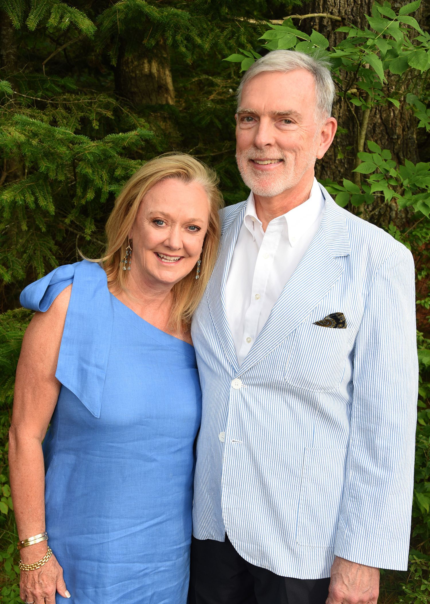 A smiling woman in a blue one-shoulder dress and a man in a blue striped blazer pose together outdoors.