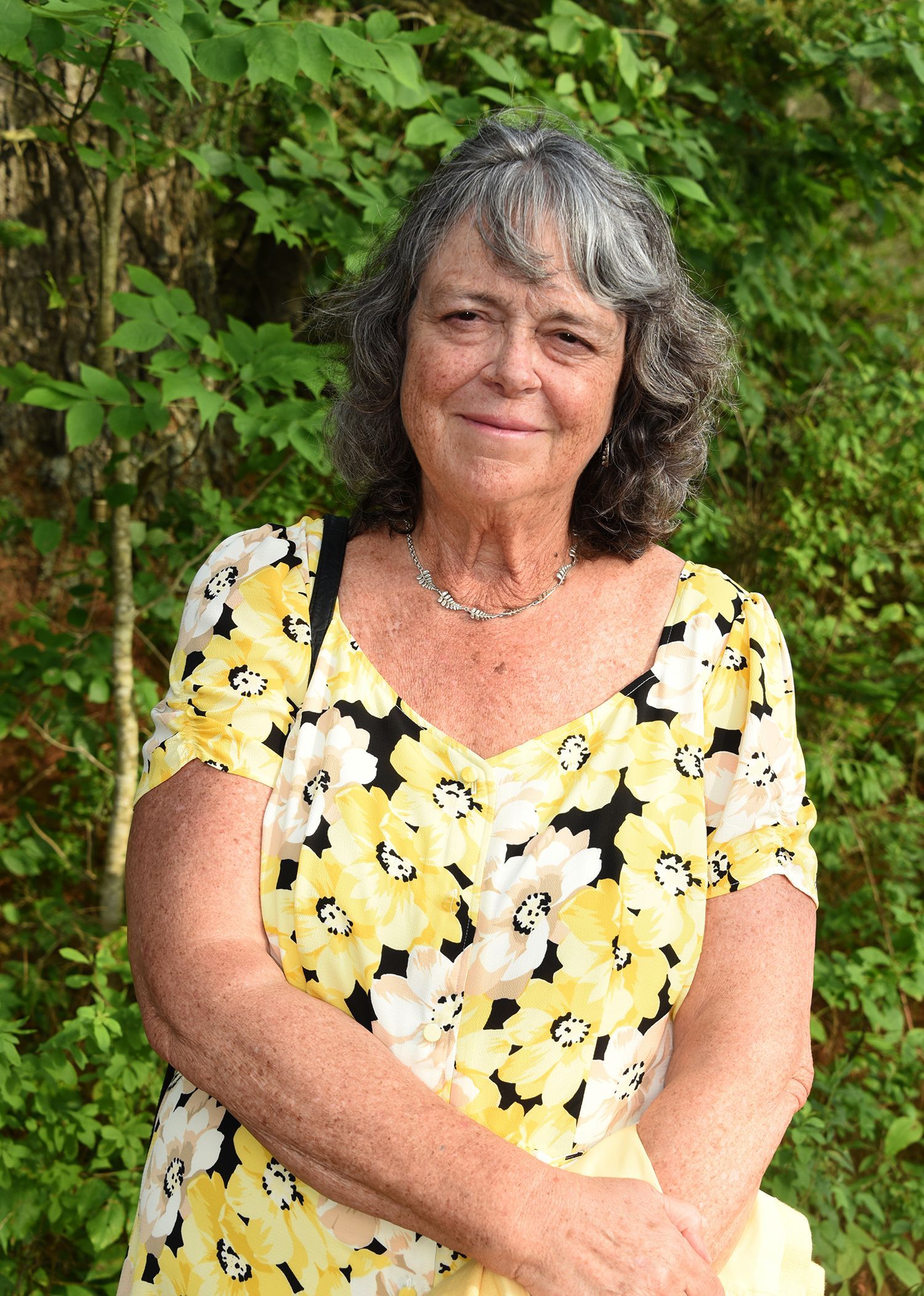 Woman with graying wavy hair and floral yellow dress smiles outdoors.