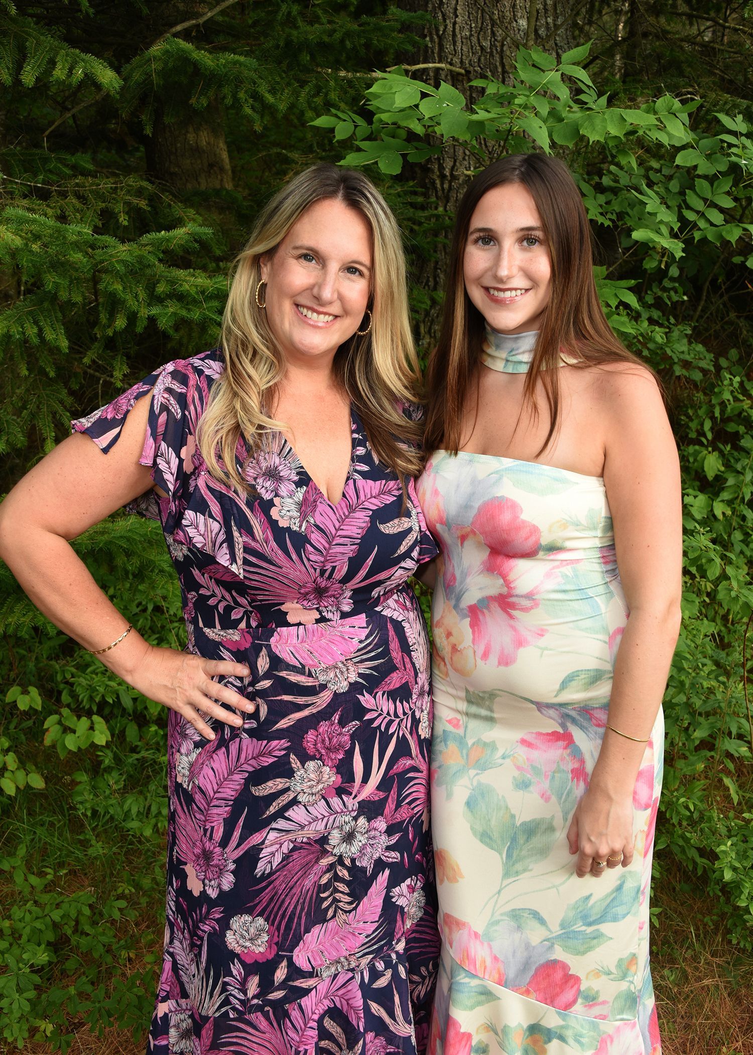 Two women smiling, posing outdoors. One in a floral dress, the other in a strapless floral gown.