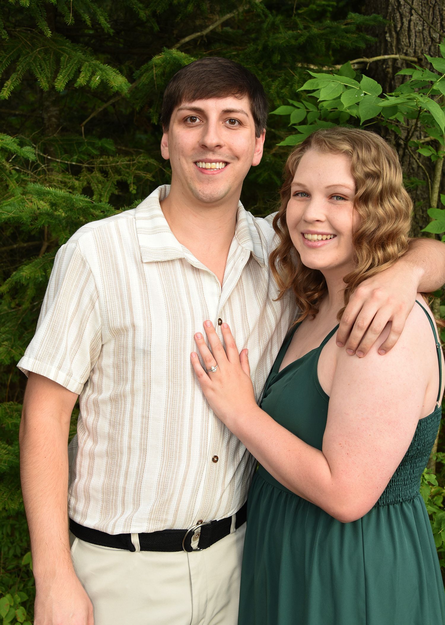 Couple posing outdoors. The man has his arm around the woman. They are smiling. The woman is wearing a green dress.