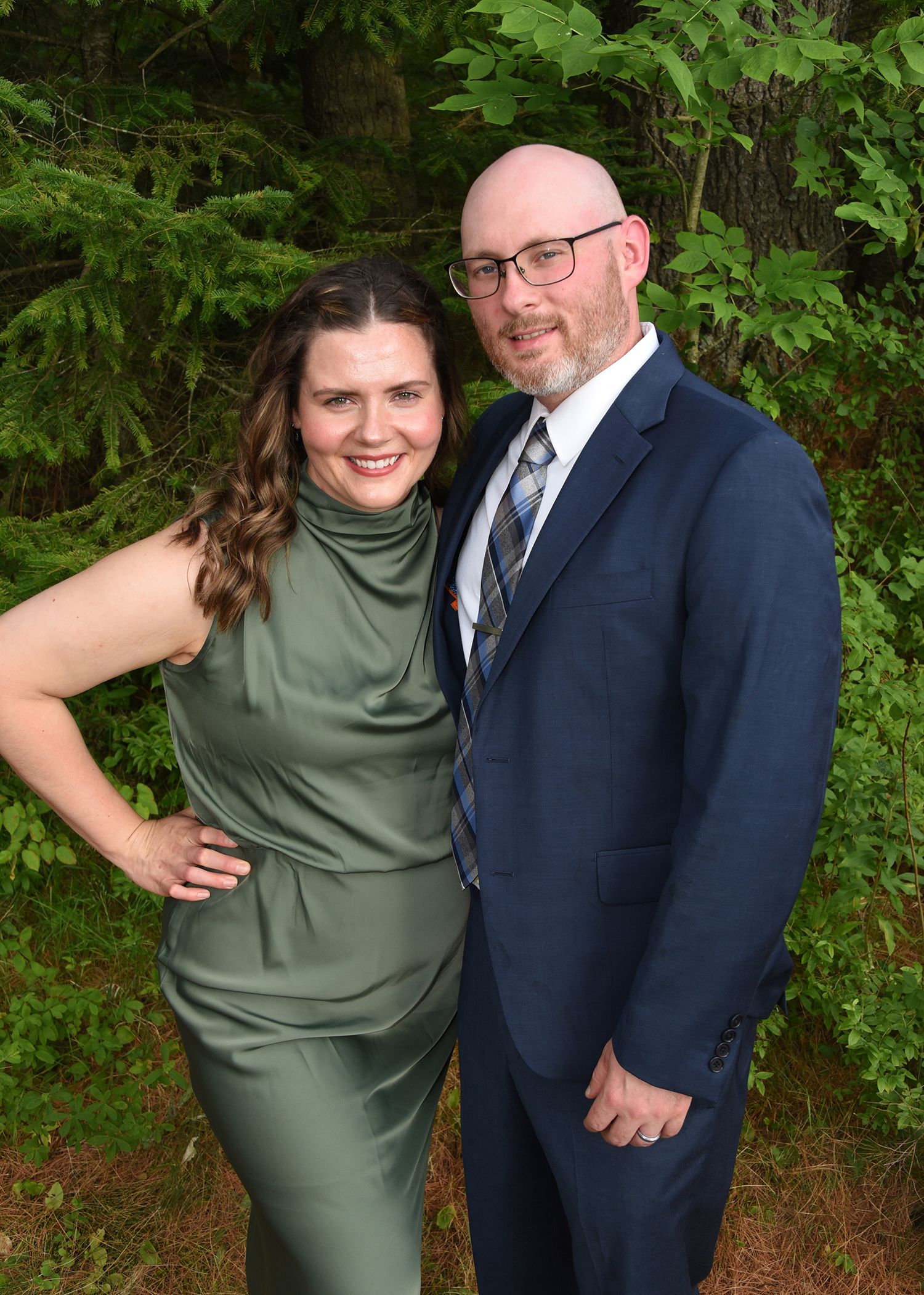 A couple poses outdoors. The woman wears a green dress, while the man dons a navy suit and tie. They are smiling.