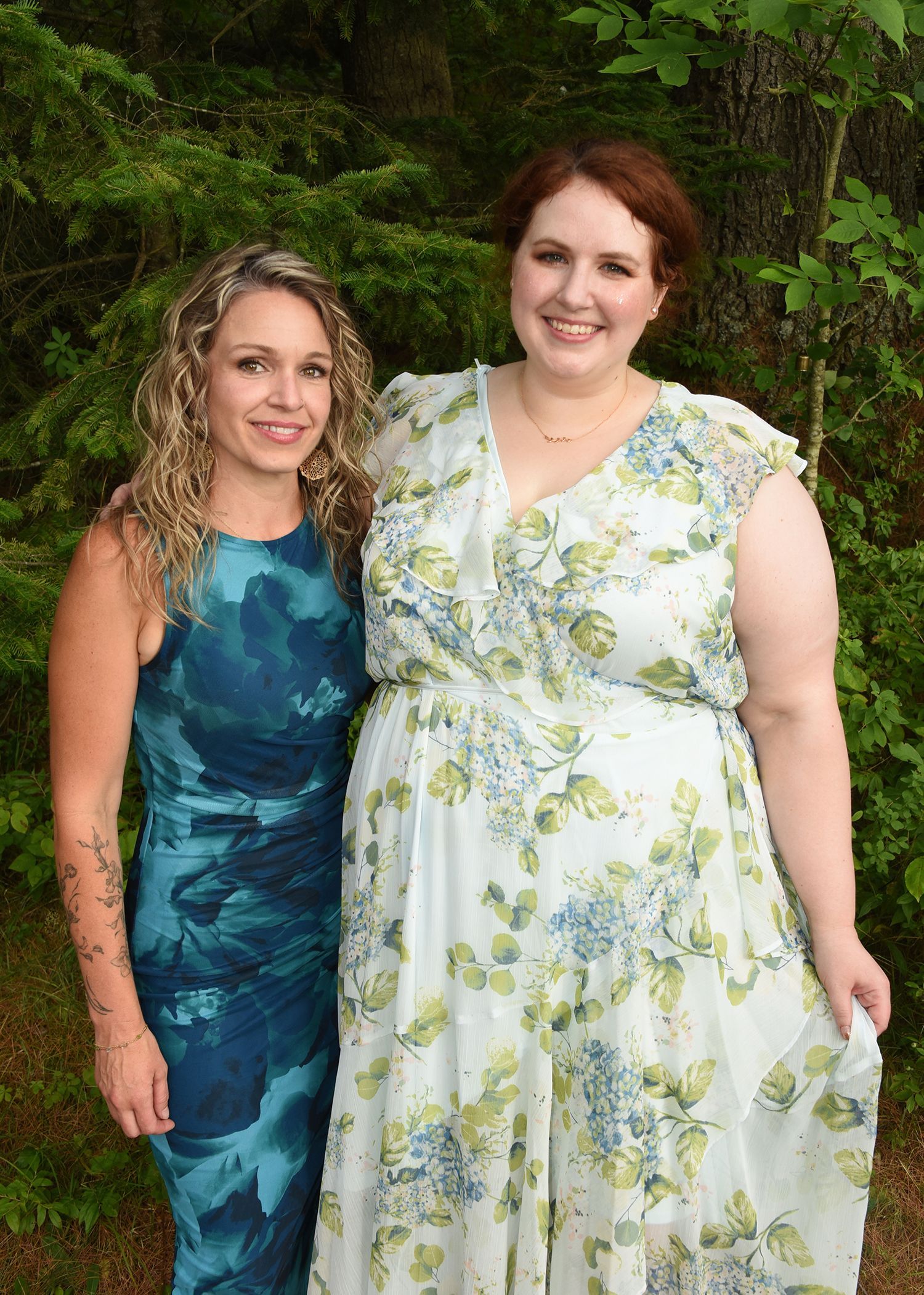 Two women smiling together in front of green foliage. One wears a blue dress, the other a floral patterned dress.