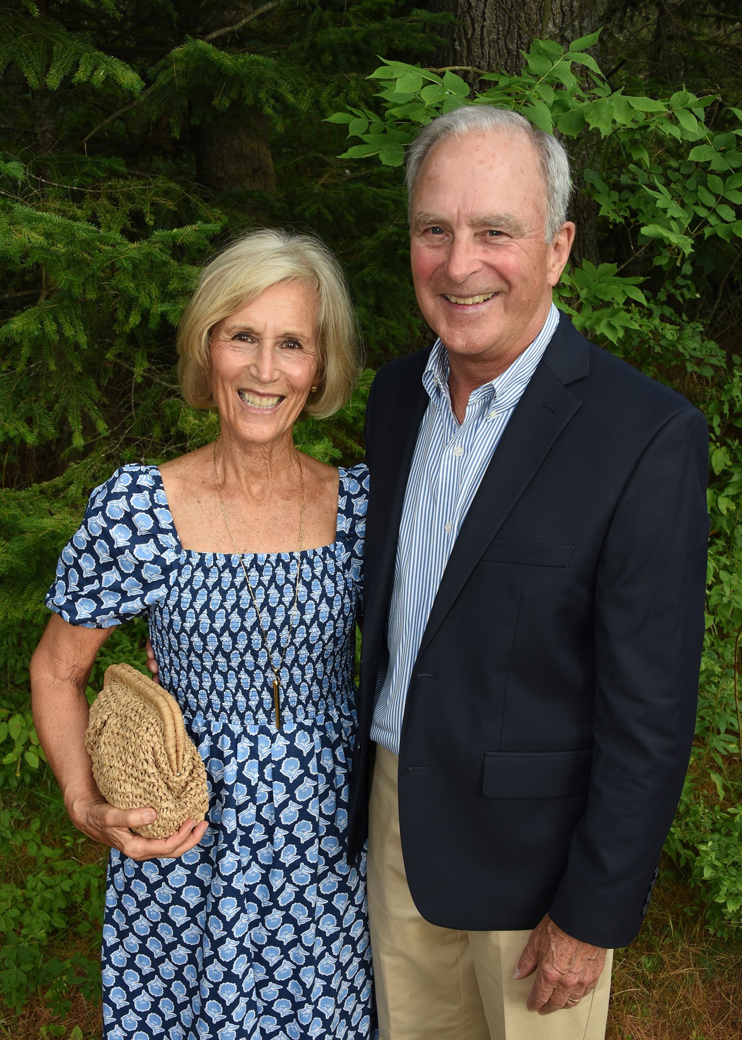 Smiling older couple posing outdoors. The woman wears a blue patterned dress and holds a woven bag. 