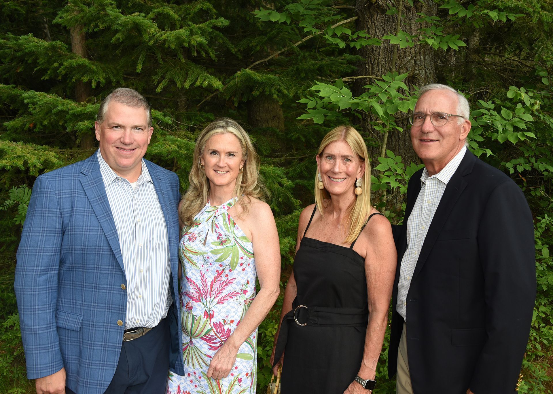 Four people posing outdoors by evergreen trees. Two women are in dresses, and two men are in suit jackets and shirts.