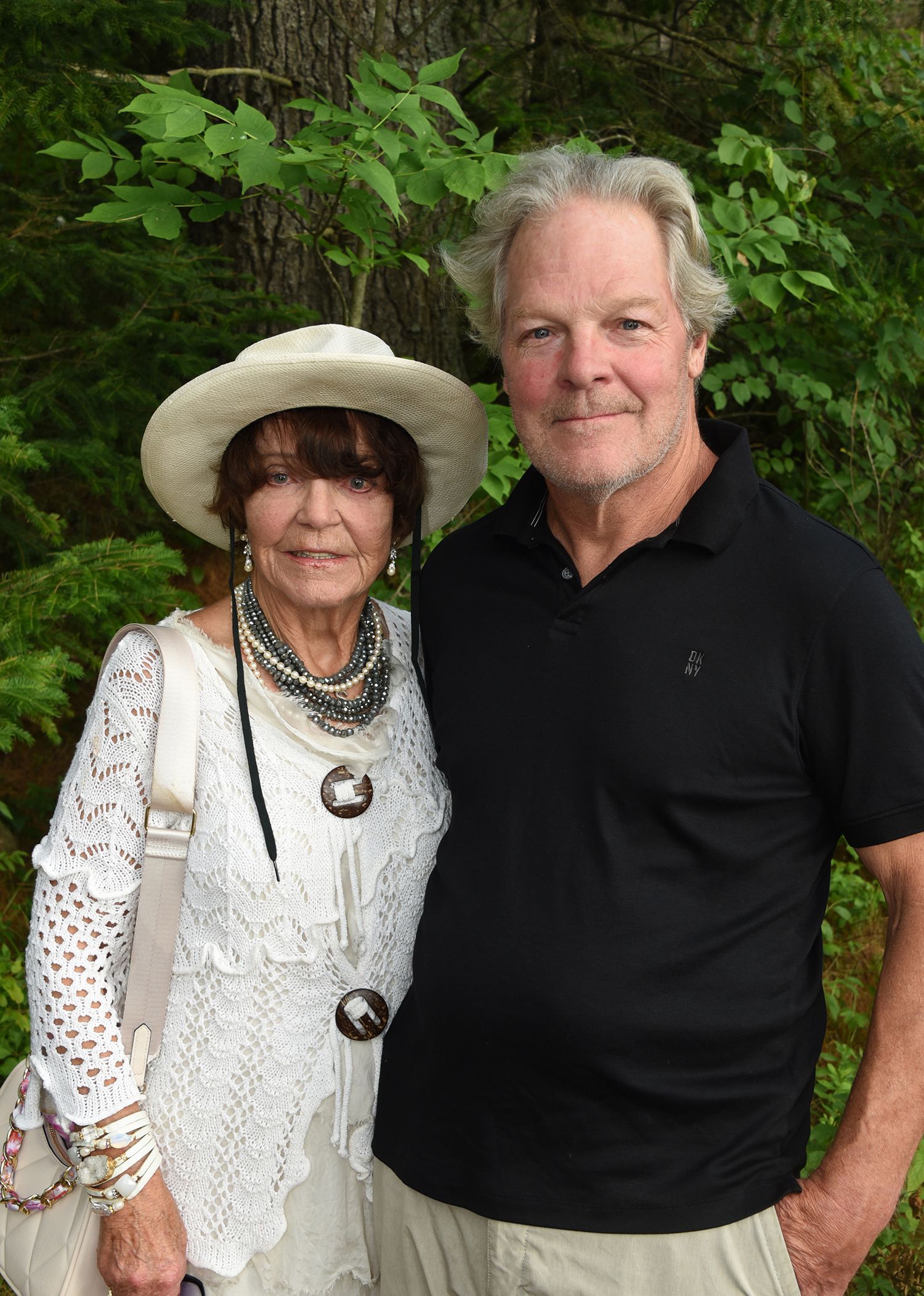 Woman in white with a sun hat and necklace, and a man in a black polo shirt, pose outside. Green foliage is behind them.