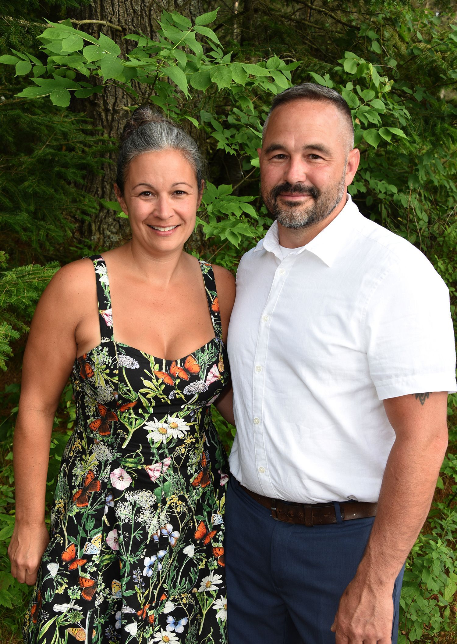 Woman in floral dress and man in white shirt, smiling, standing outdoors. Behind them is green foliage.