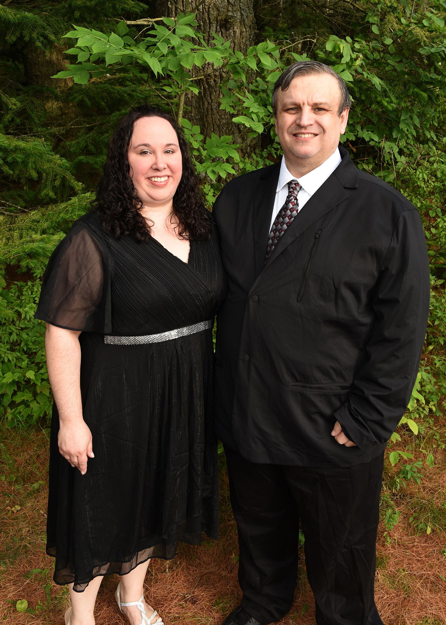 A smiling woman and man stand close together outdoors. The woman wears a black dress, and the man wears a black suit and tie.