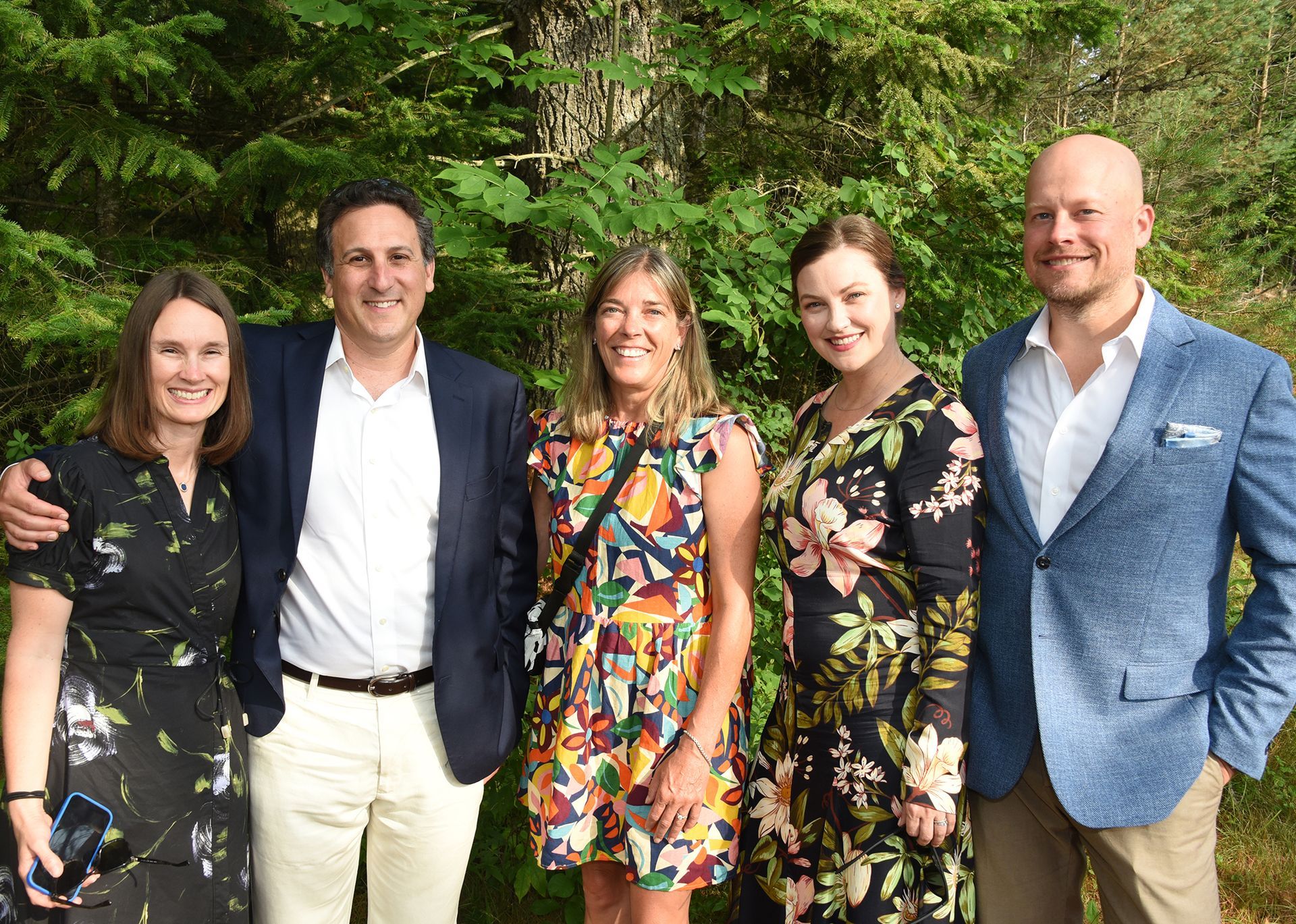 Five people smiling, posed outdoors in front of green foliage. One man has his arm around a woman.