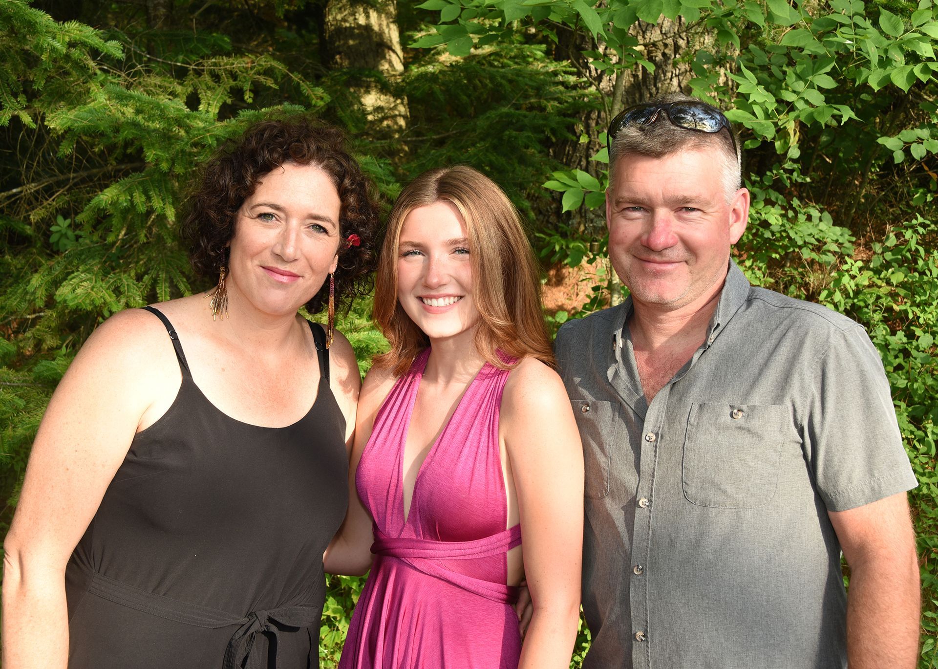 Woman in black dress, young woman in pink dress, and man in grey shirt smiling in front of foliage.