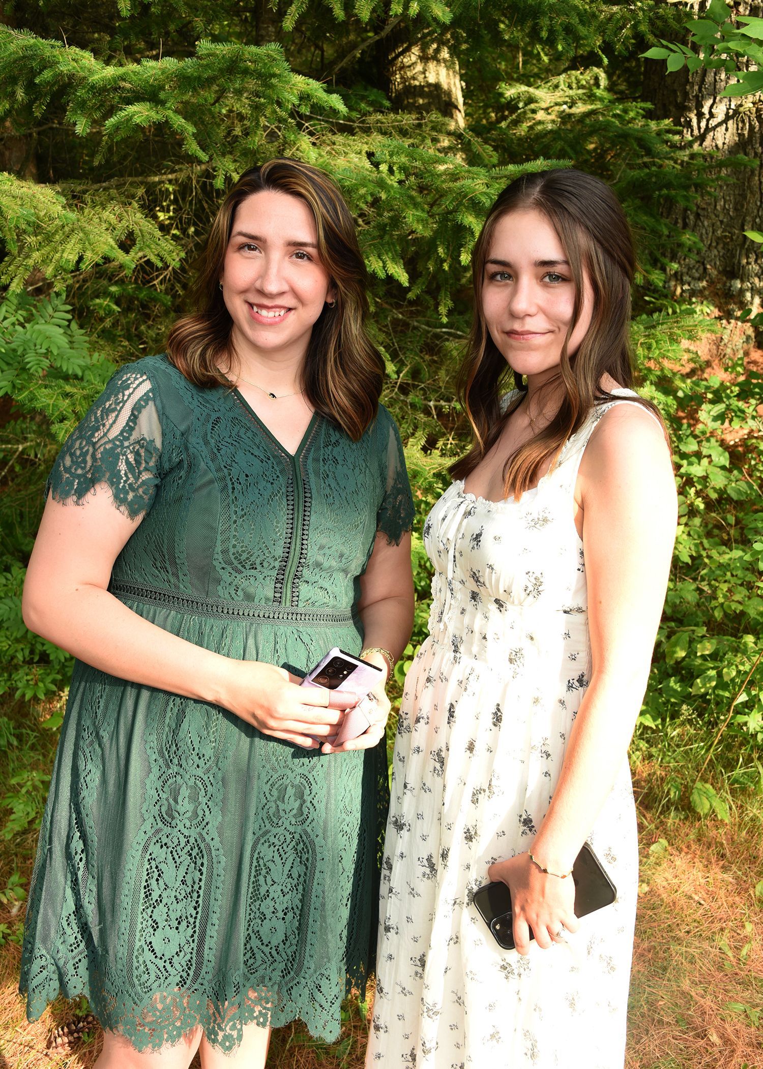 Two women smiling in front of lush greenery. One wears a green lace dress, the other a white floral dress.
