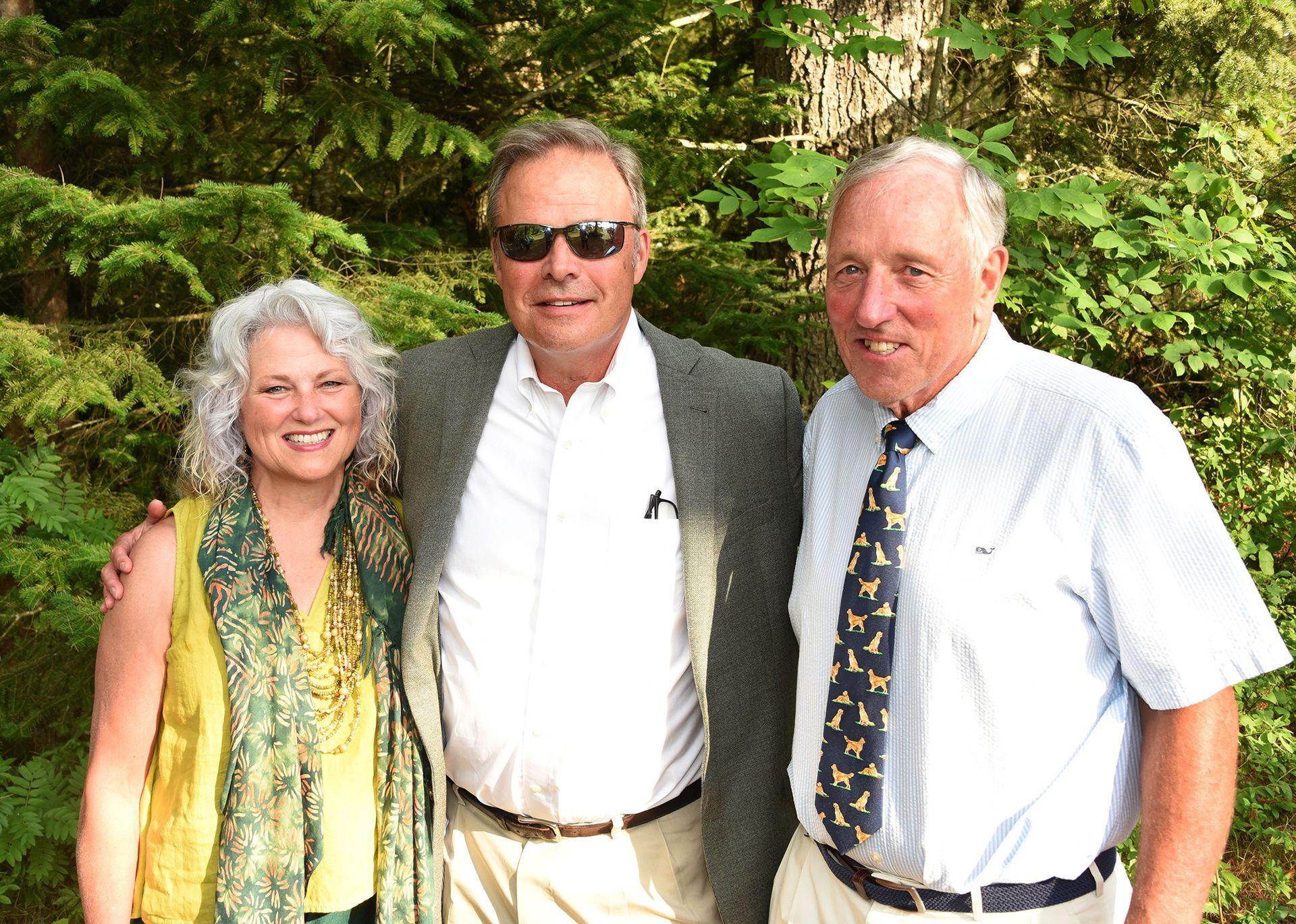 Three people posing outdoors, smiling. A woman with curly grey hair and a yellow scarf stands to the left.