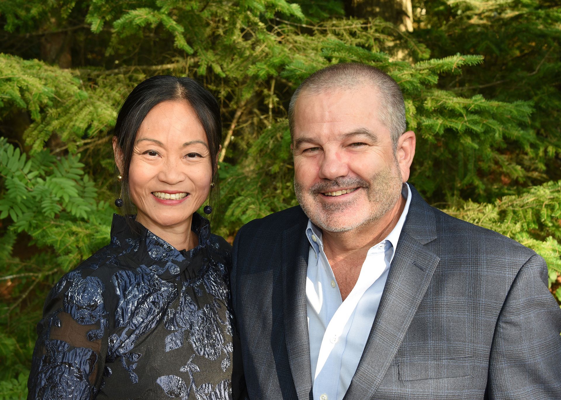 Woman in black floral top and man in gray jacket smiling in front of green foliage.