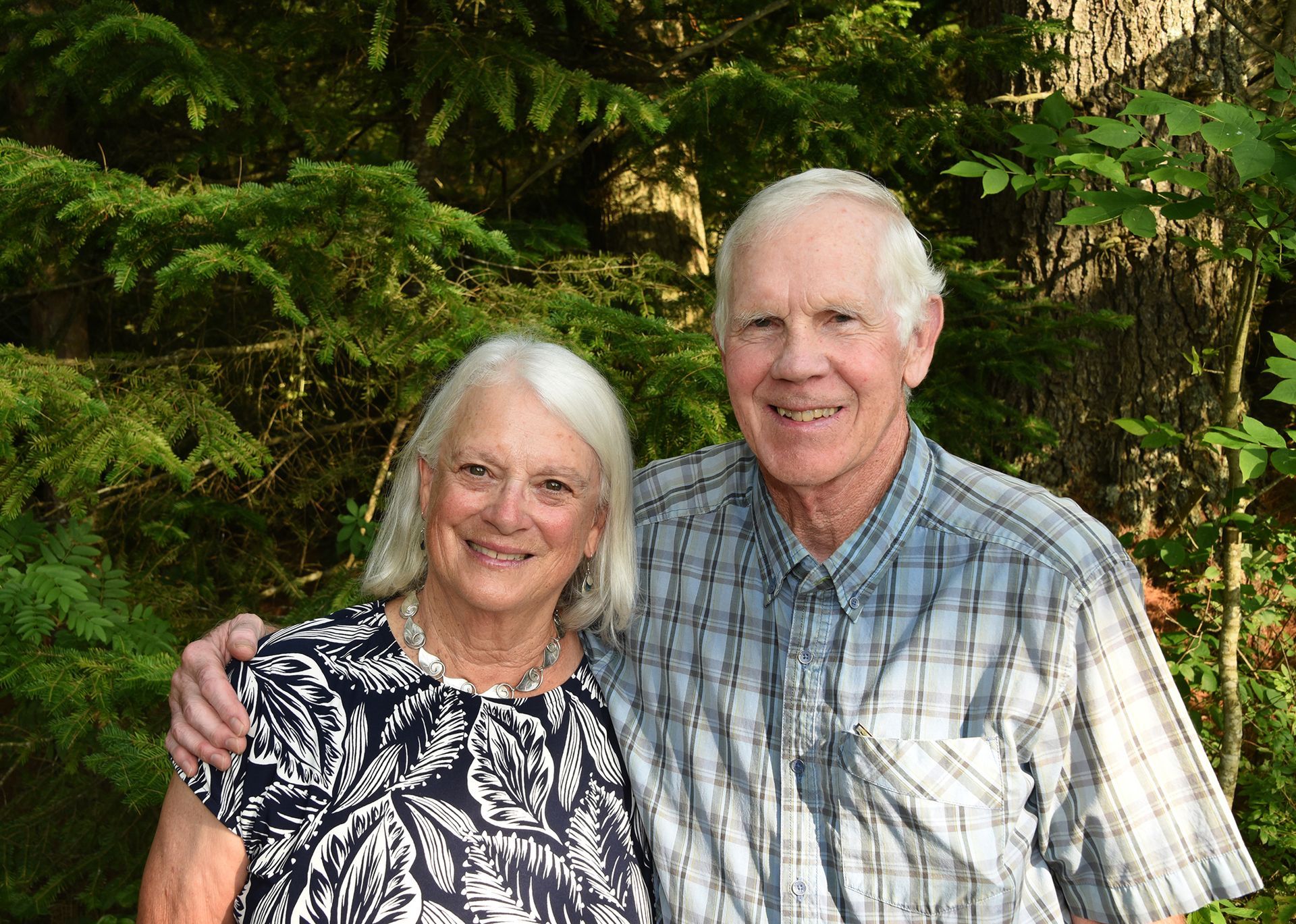 Elderly couple smiling, posing together in front of a green, leafy backdrop. Man has his arm around the woman.
