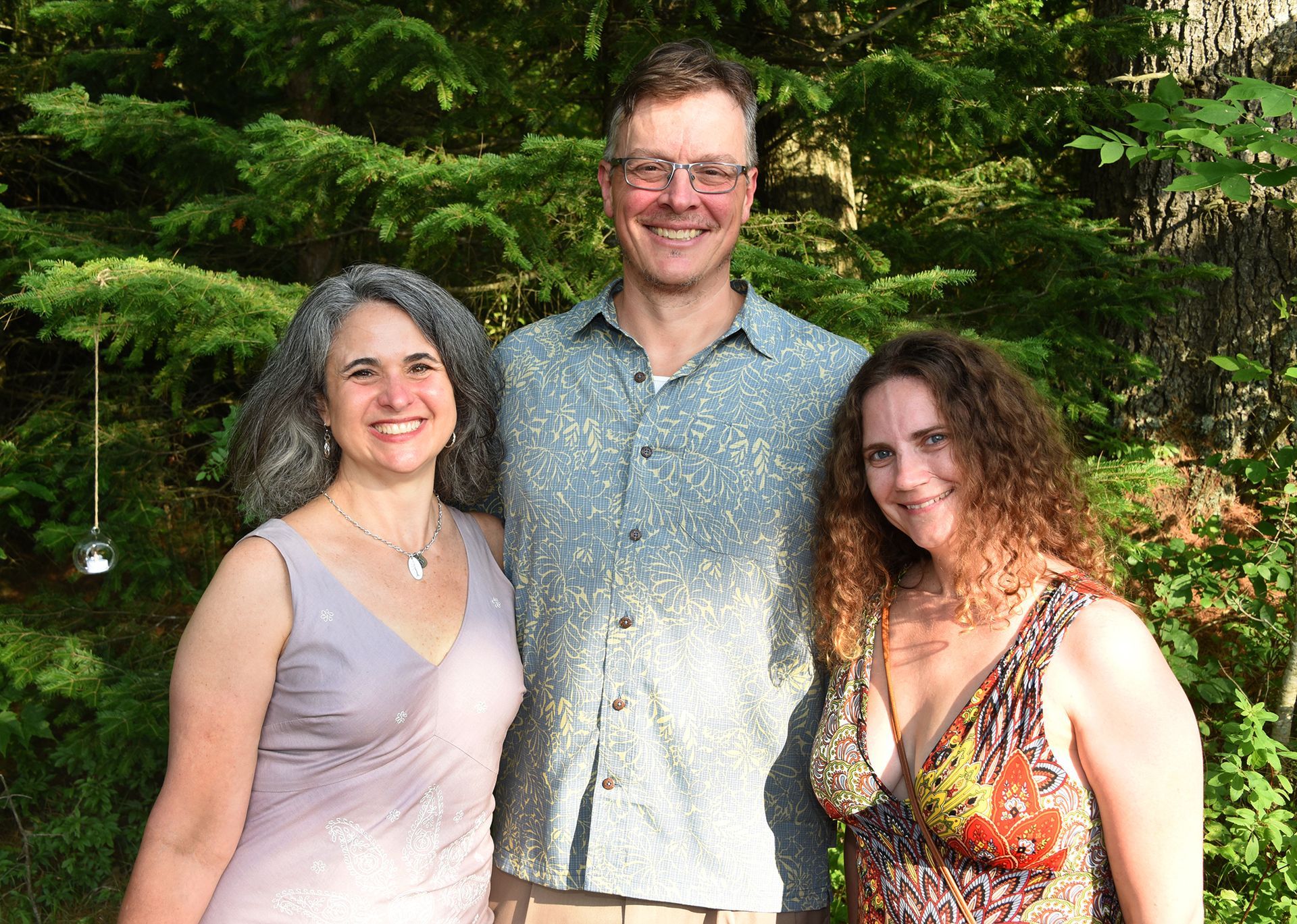 Three smiling adults pose outdoors. A man in glasses stands between two women, all in front of a green backdrop of trees.